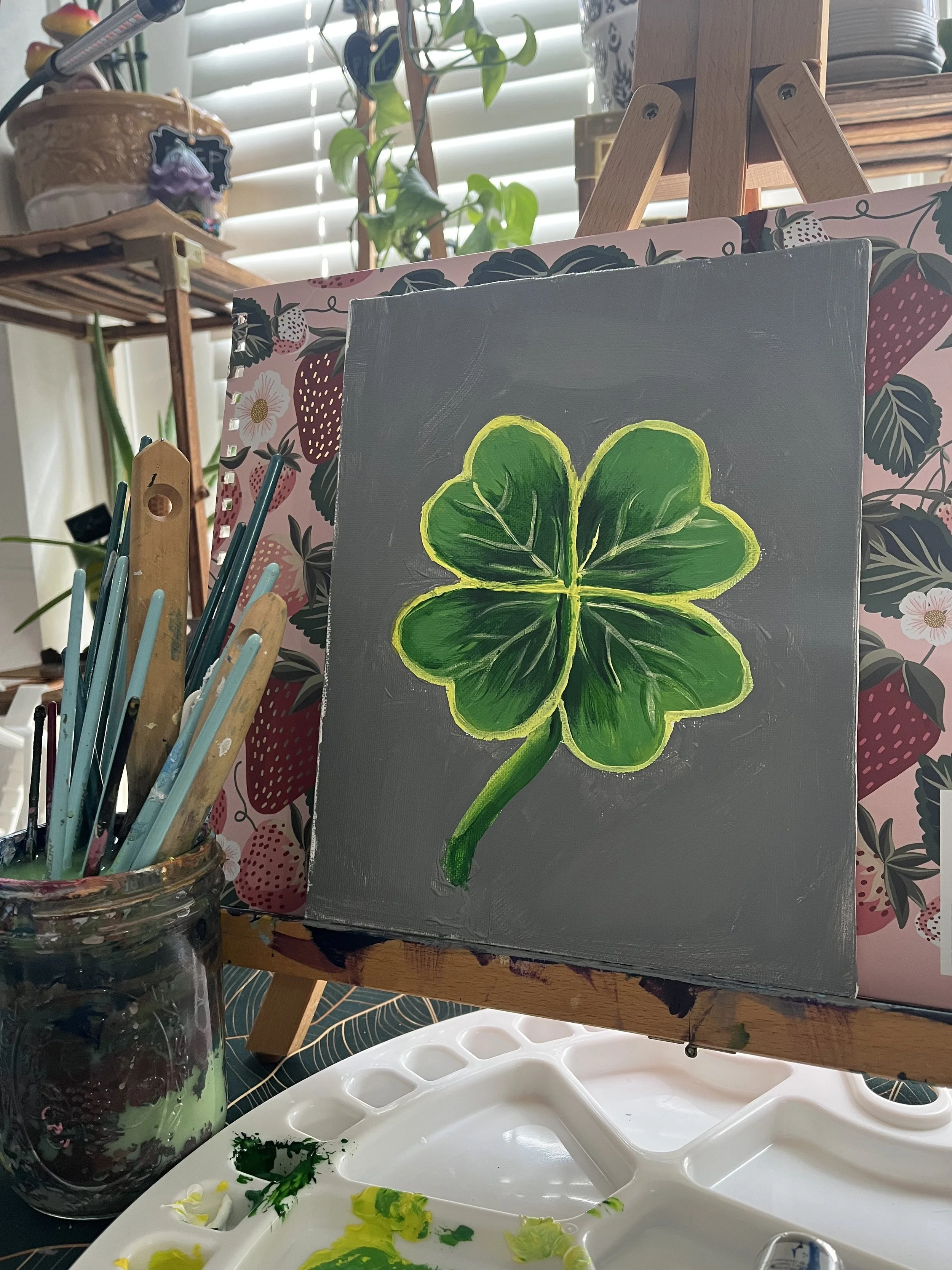 A painting of a green four-leaf clover on a gray canvas, set on an easel in an artist's workspace with painting brushes in a jar and a palette.
