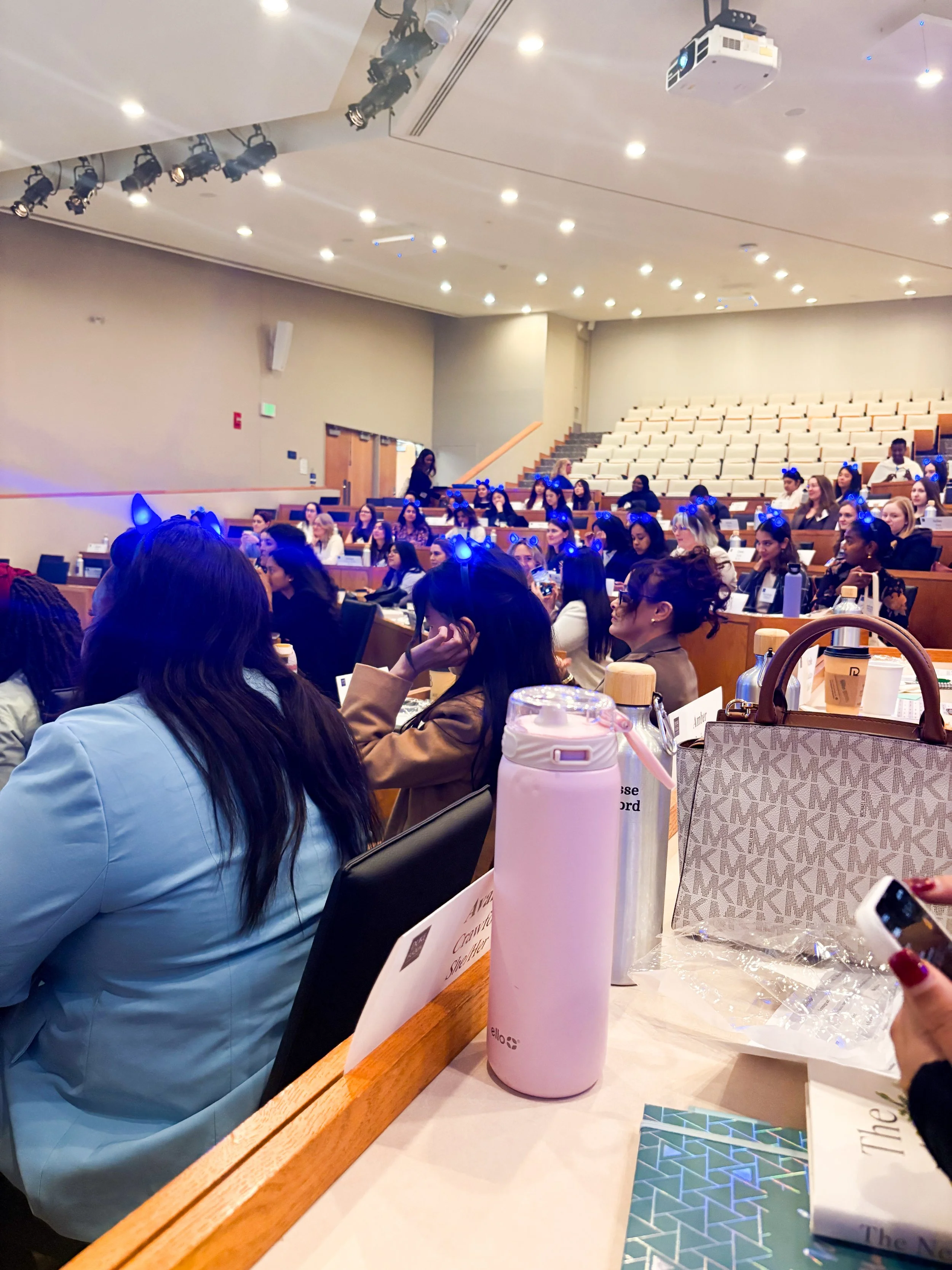 Women attending a professional leadership conference in a large auditorium with a speaker presenting at the front of the room.