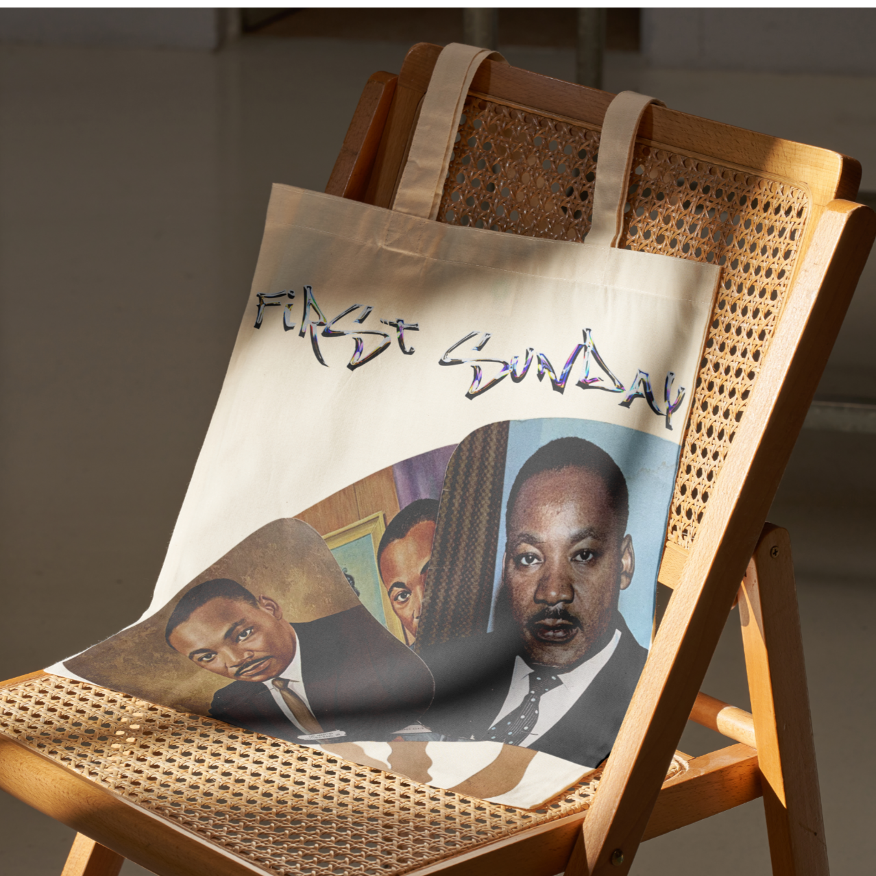 A tote bag with the words "First Sun" hanging on a wooden chair with a woven cane back, featuring images of a man in a suit.