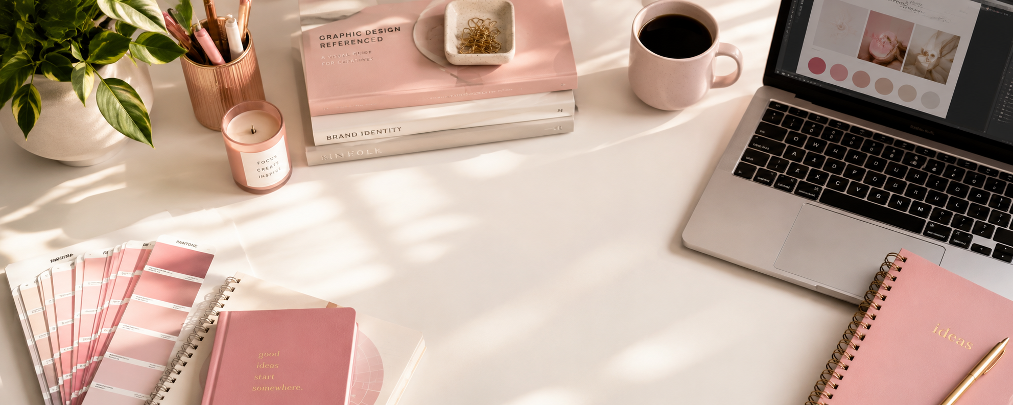 A flat lay of a creative workspace with a white desk, Pink notebooks, a color swatch chart, a potted plant, a pink pen holder with pens and markers, a pink candle, a stack of books titled 'Graphic Design Referenced' and 'Brand Identity', a small dish with gold paper clips, a pink coffee mug, and an open laptop displaying design software, with natural light and shadows.