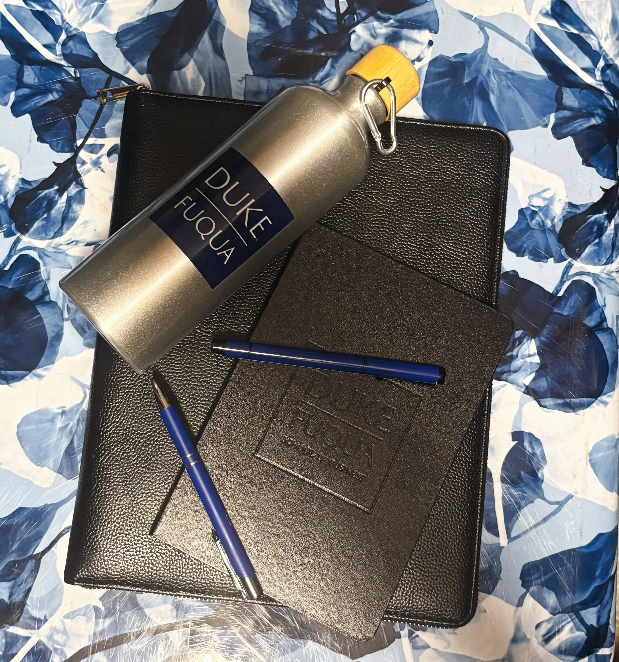 Duke Fuqua branded notebook with pens and water bottle arranged on a patterned surface representing graduate school materials.