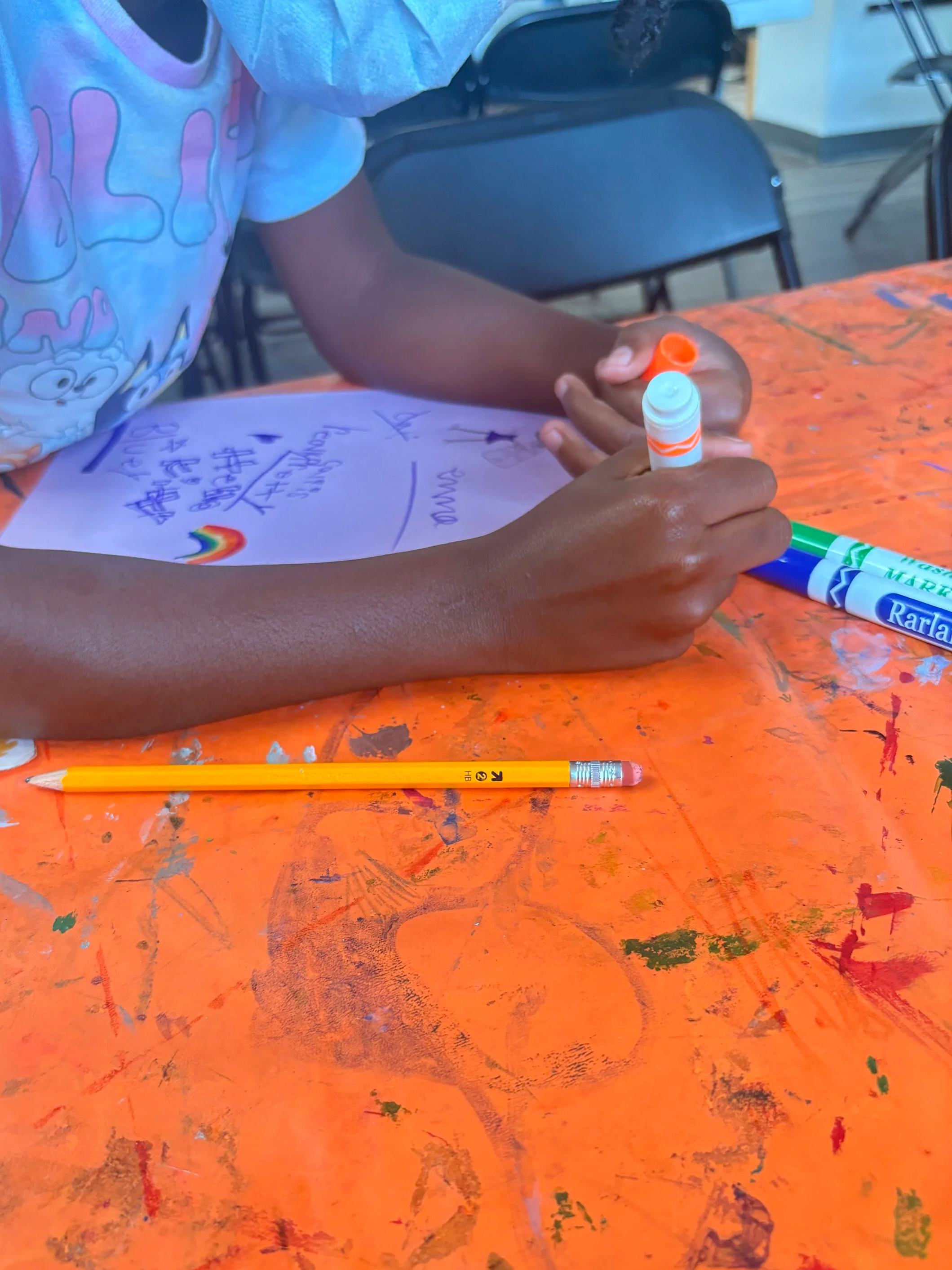 A person writing a message on a piece of paper with colorful markers, sitting at an orange table that has paint stains and scribbles.