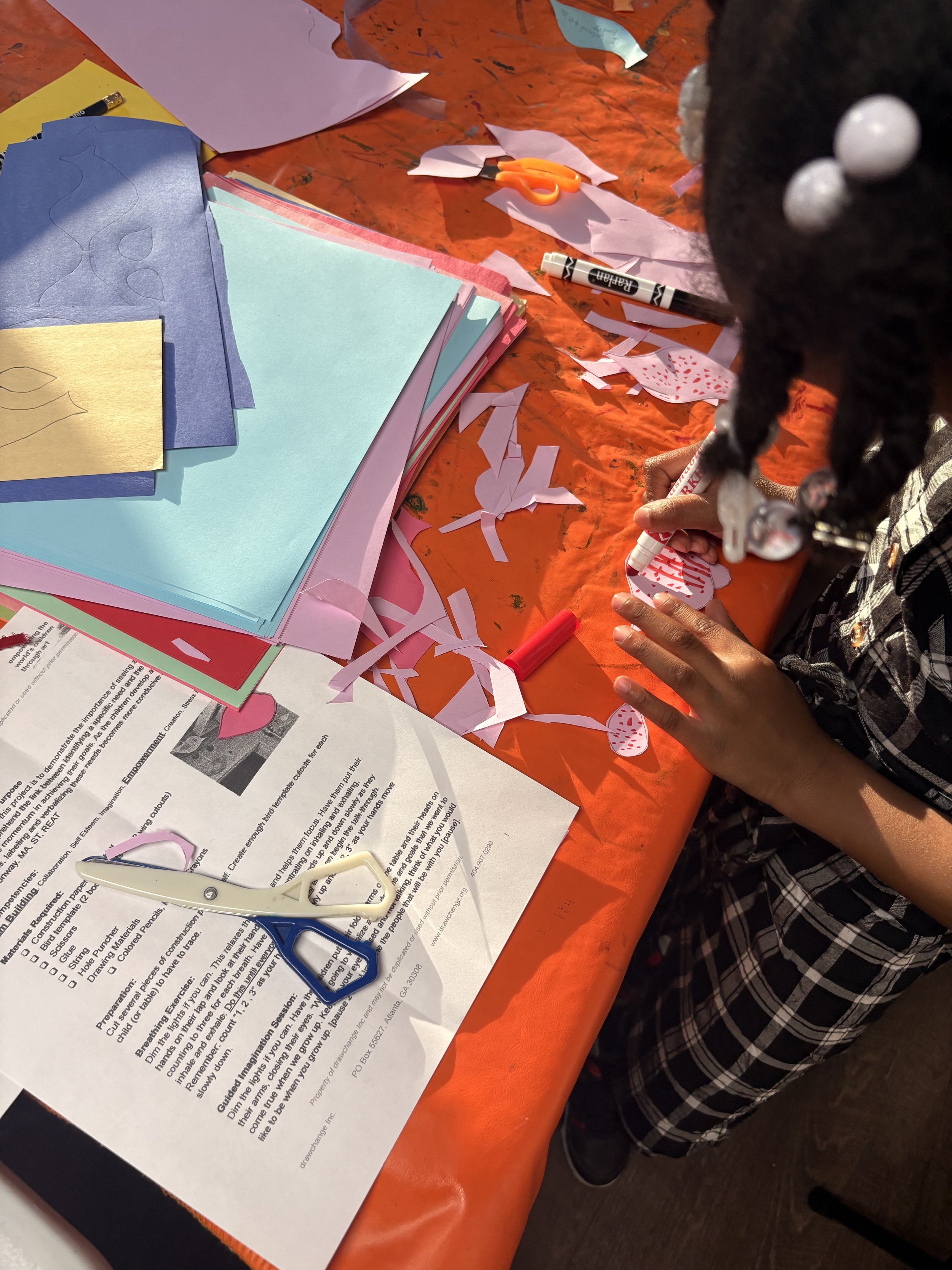 Child working on a colorful craft project with scissors, markers, and paper materials spread across a table.