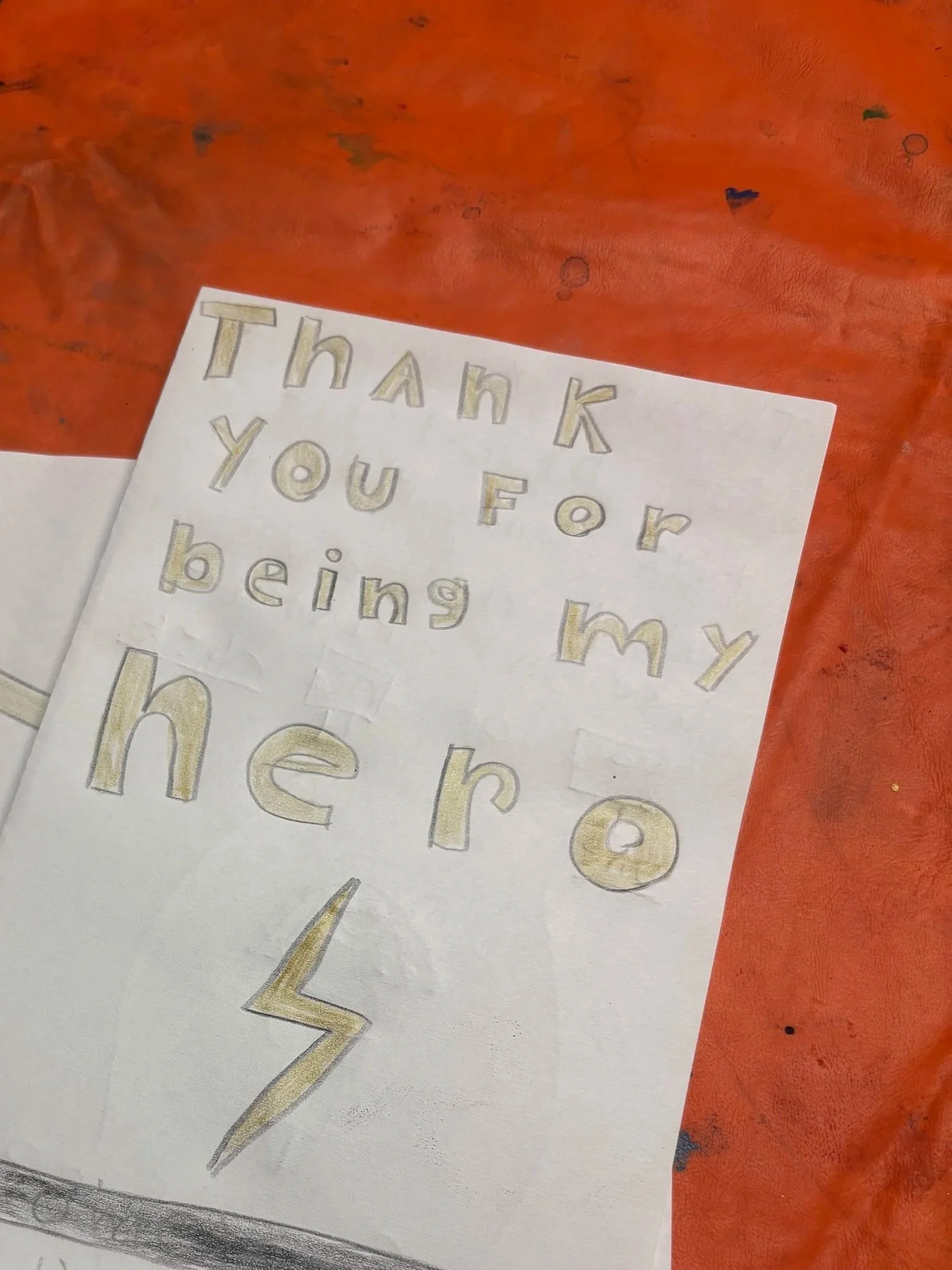 A handwritten note on white paper with the words 'Thank you for being my hero' written in large, gold and gray outlined letters. The paper is placed on a textured, orange surface.