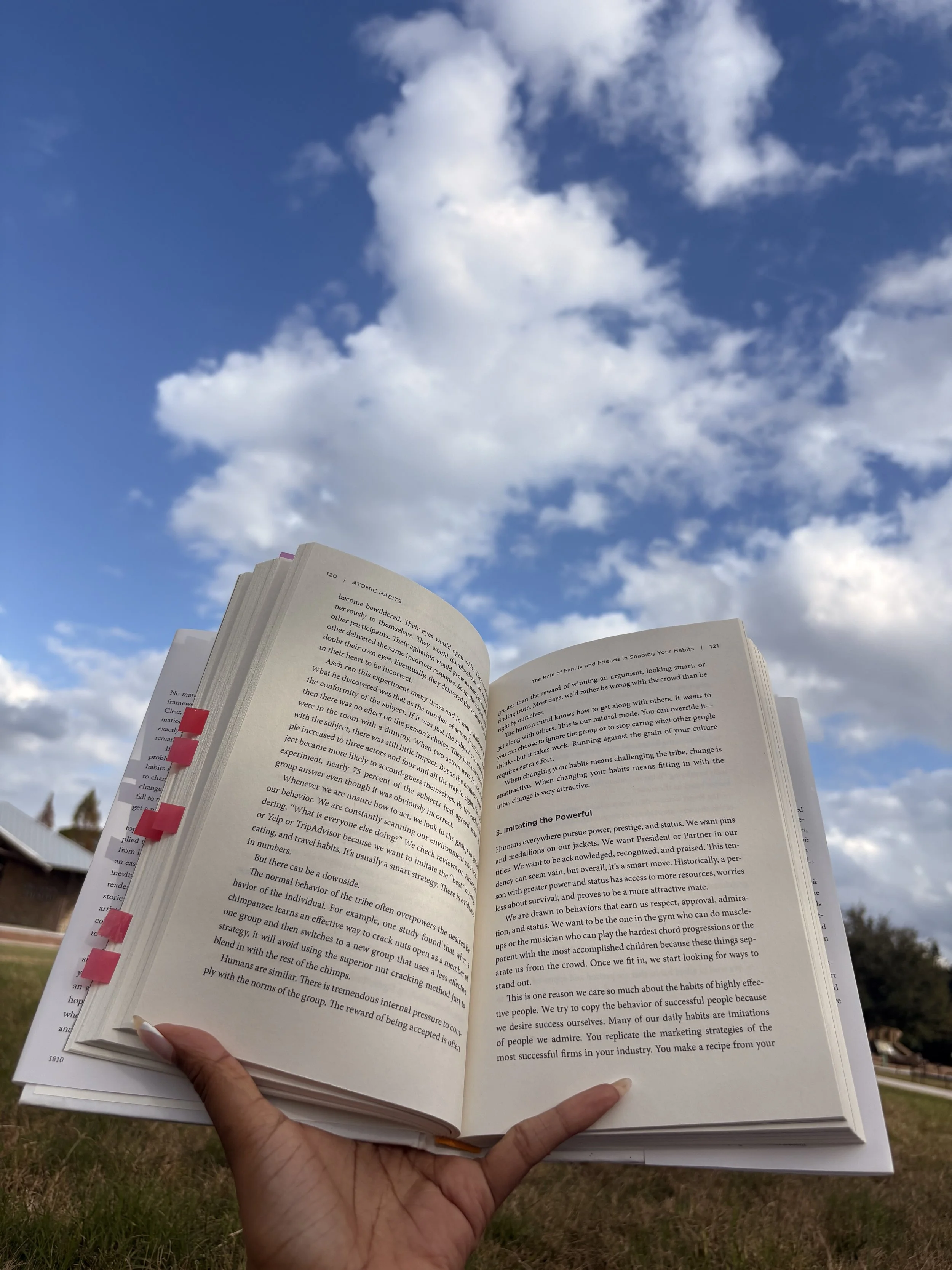 Person holding an open book outdoors against a bright blue sky with clouds, representing reading and personal development.