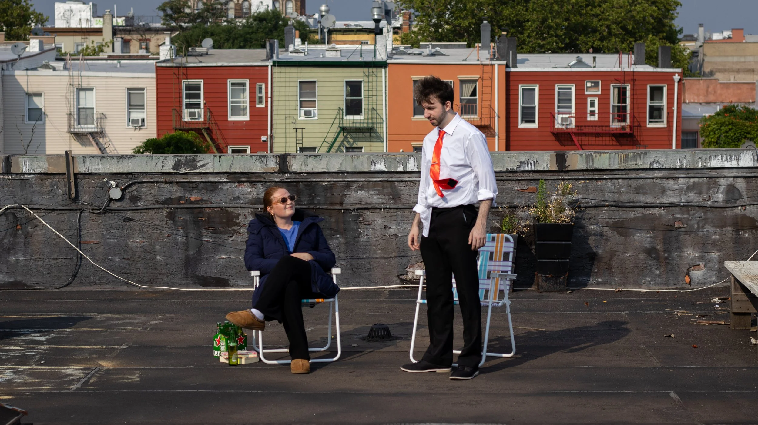 A man in a white shirt and black pants with a red tie stands on a rooftop, talking to a woman seated in a camping chair, wearing sunglasses and a dark jacket. The woman has a pack of Heineken beer bottles on the ground beside her. In the background,
