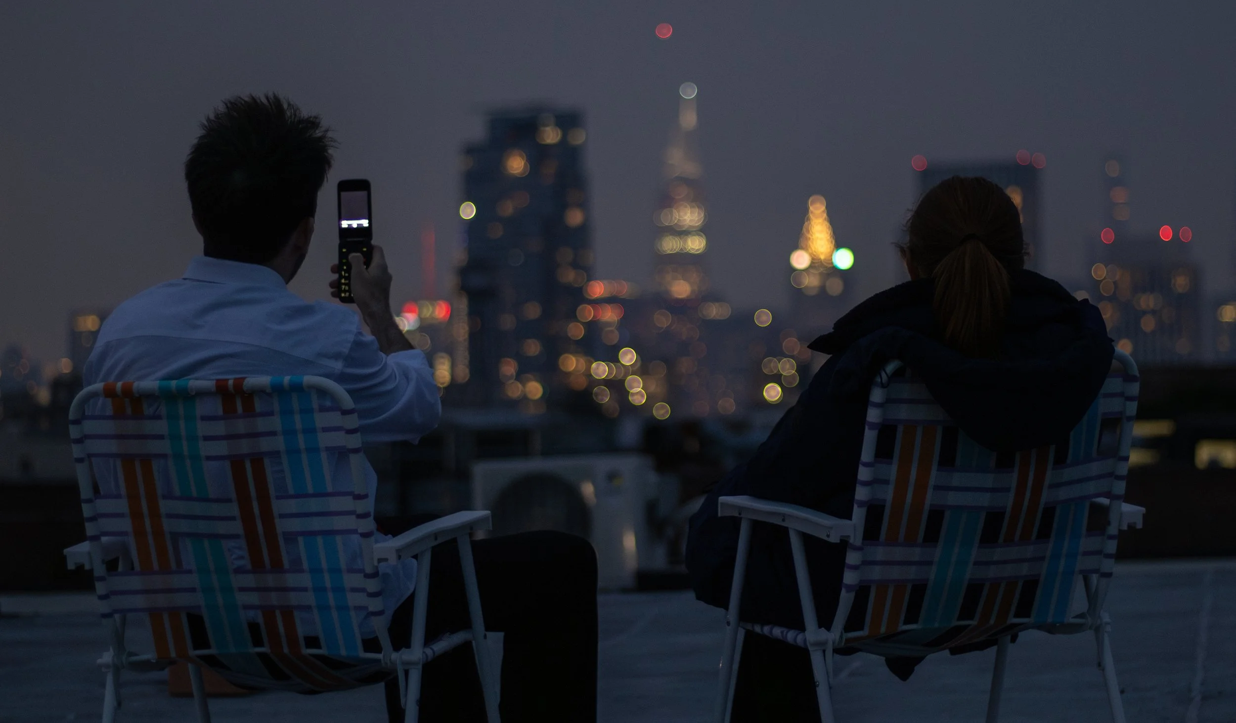 Two people sitting on chairs on a rooftop at night, one taking a photo of a city skyline with a phone, city lights and tall buildings blurred in the background.