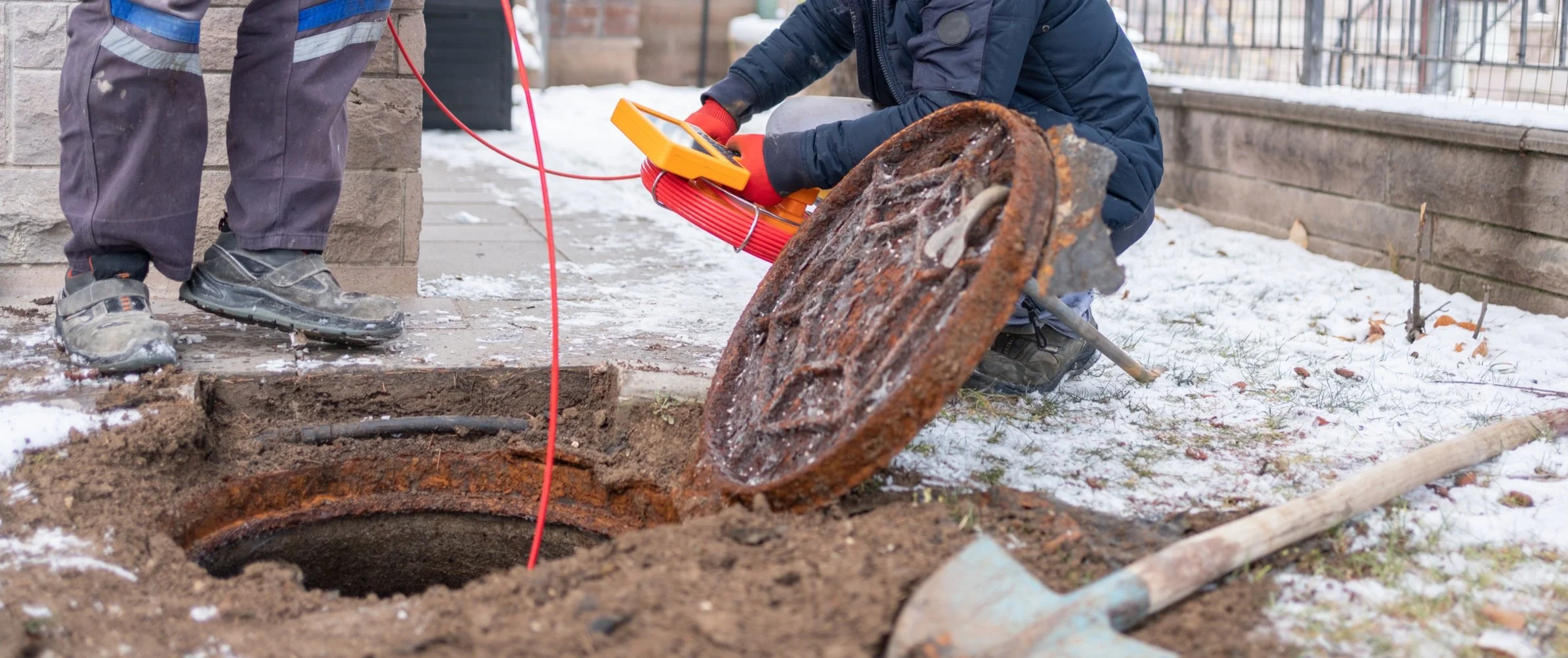Workers repairing a buried underground utility or sewer access point outdoors, with one person using testing equipment and the other standing nearby.