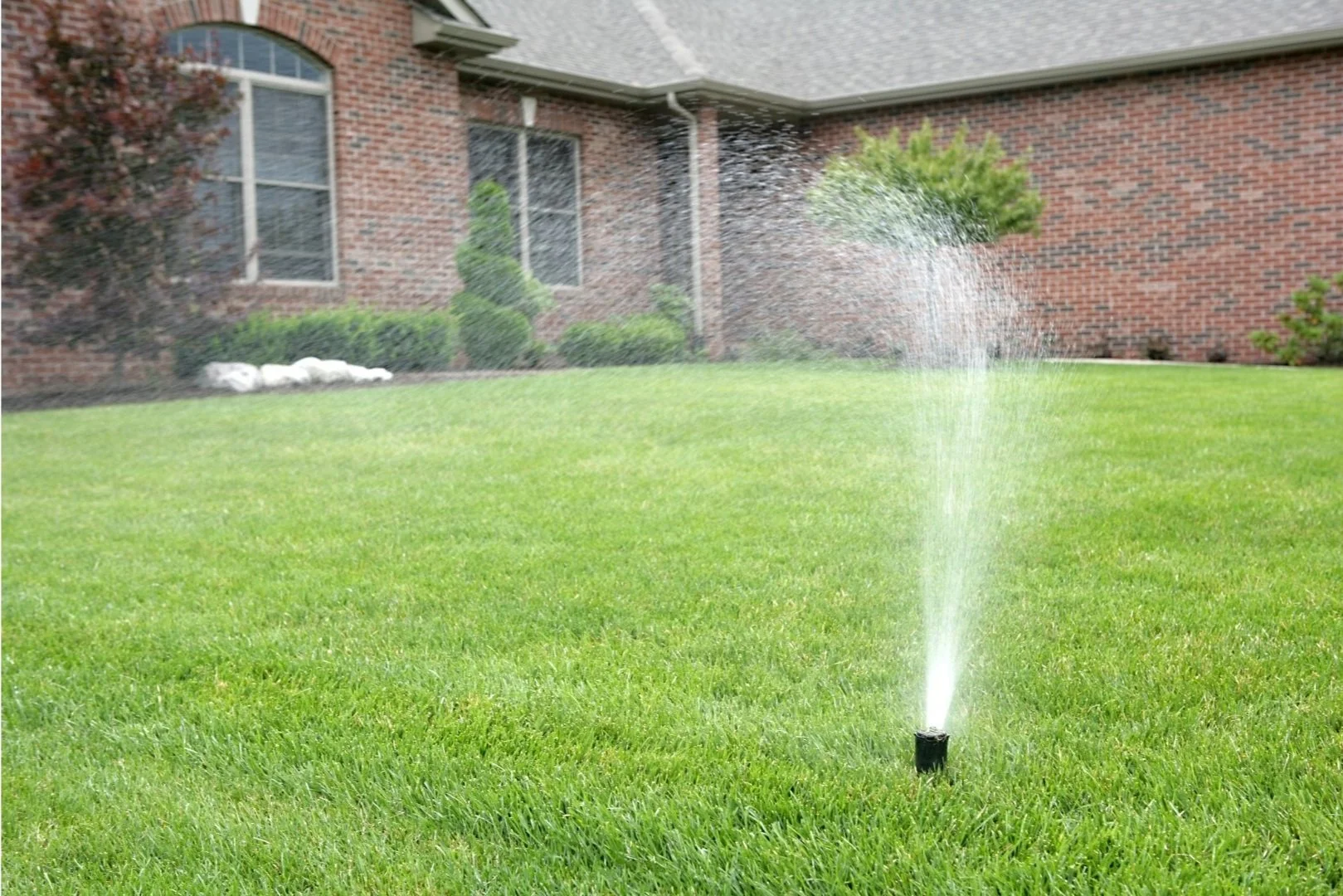 Aquamen Yorkshire Sprinkler watering system installed on green lawn in front of a brick house with windows and bushes.