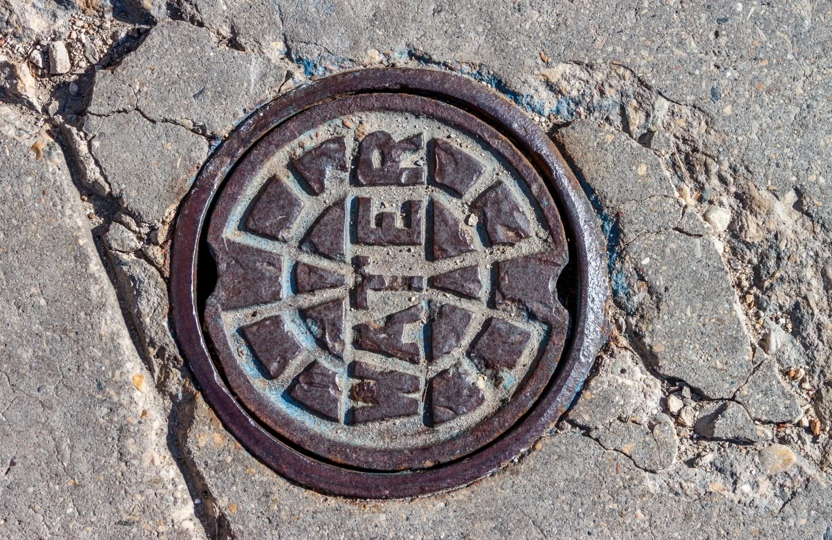 A round manhole cover with a geometric pattern in the center, surrounded by a concrete sidewalk with cracks.