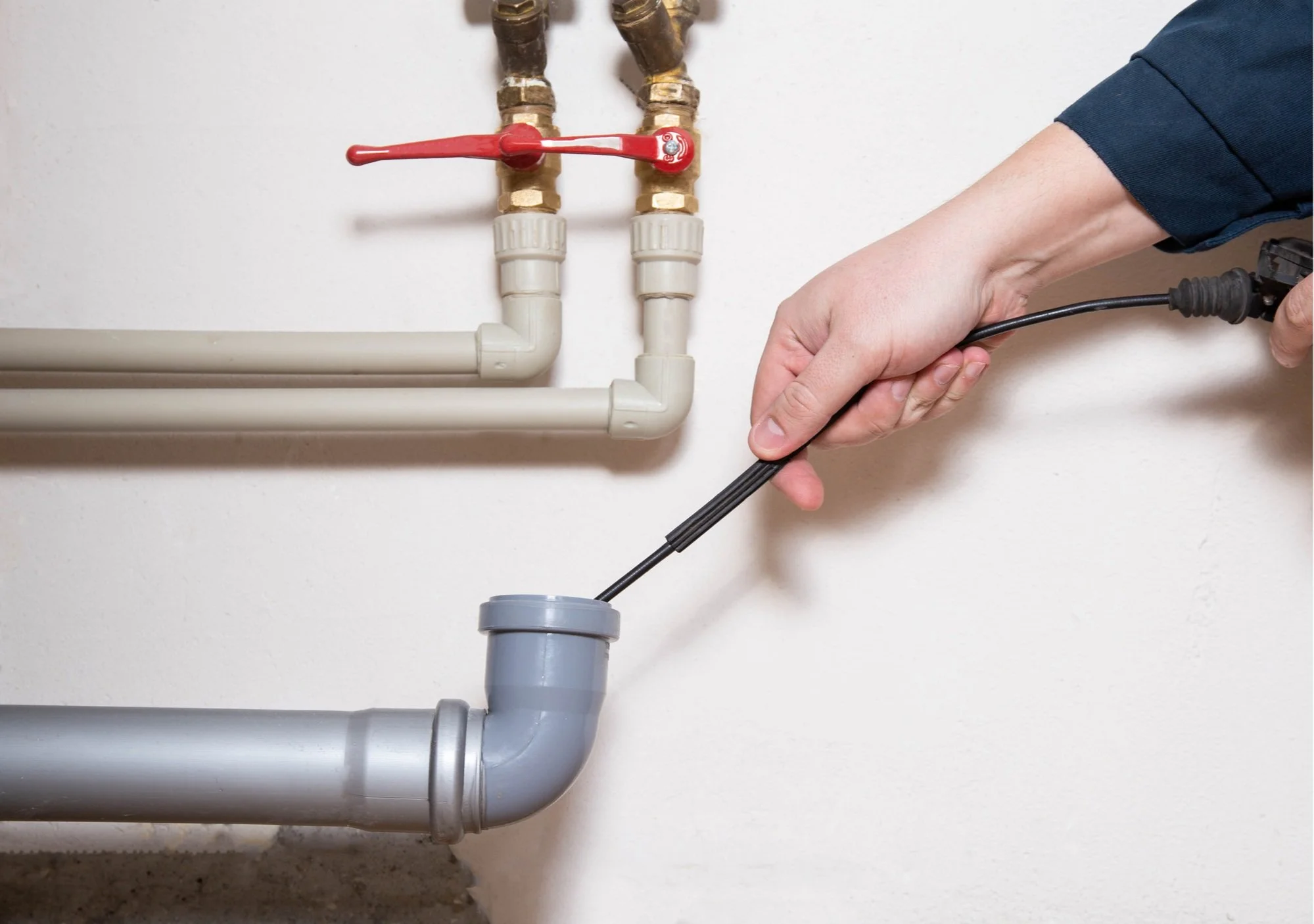 A person inspecting or repairing plumbing pipes with a screwdriver in a utility room, showing copper, PVC, and gray drainage pipes on a white wall.