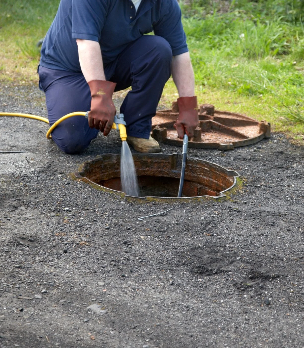 A person wearing brown gloves is using a tool to remove a manhole cover from the ground. The person is kneeling on the dirt next to the open manhole, with another removed cover placed nearby. The surrounding is grassy.