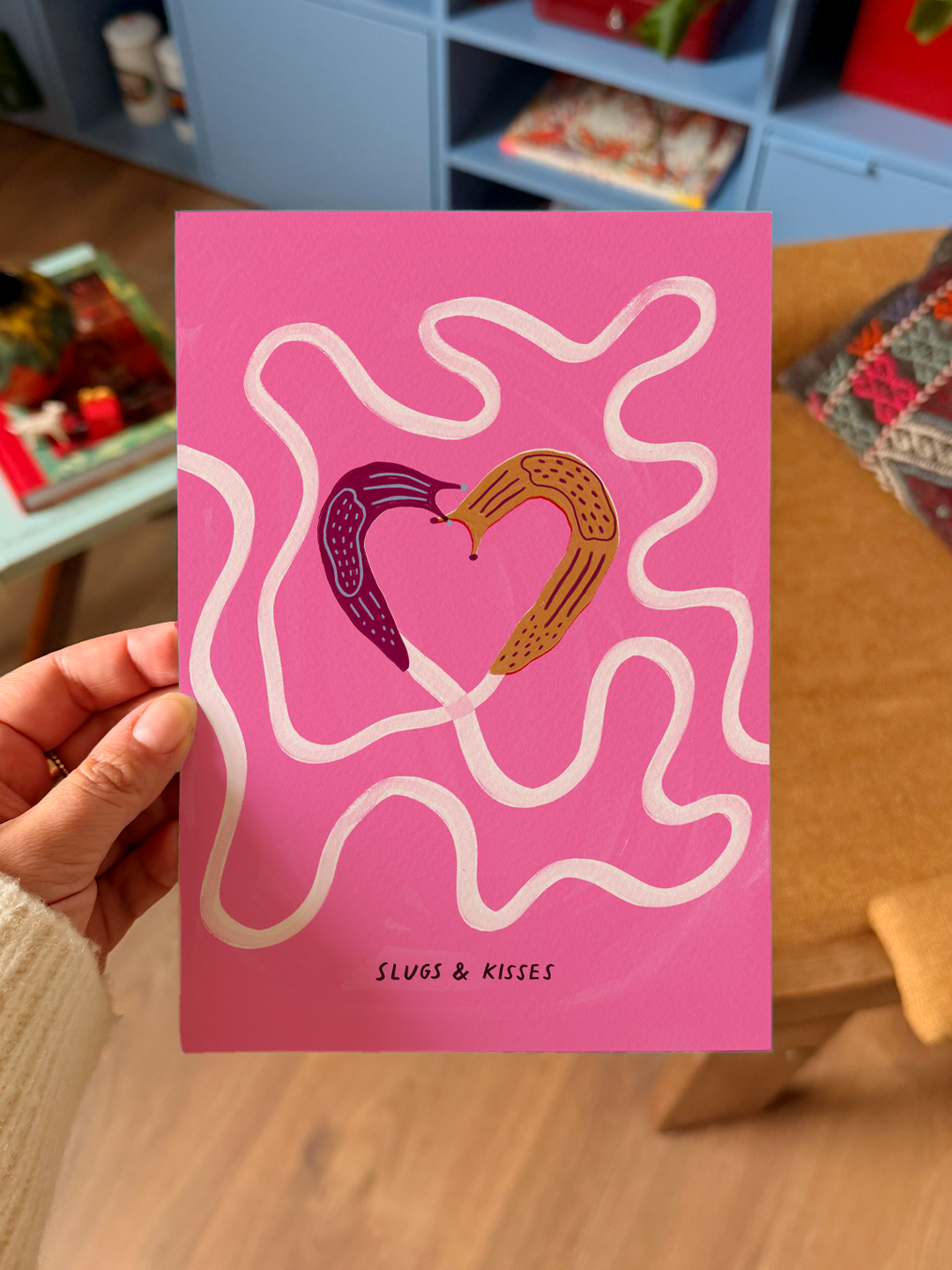 A pink greeting card with an illustration of two slugs forming a heart shape, surrounded by wavy white lines. The text on the card reads 'SLUGS & KISSES.'