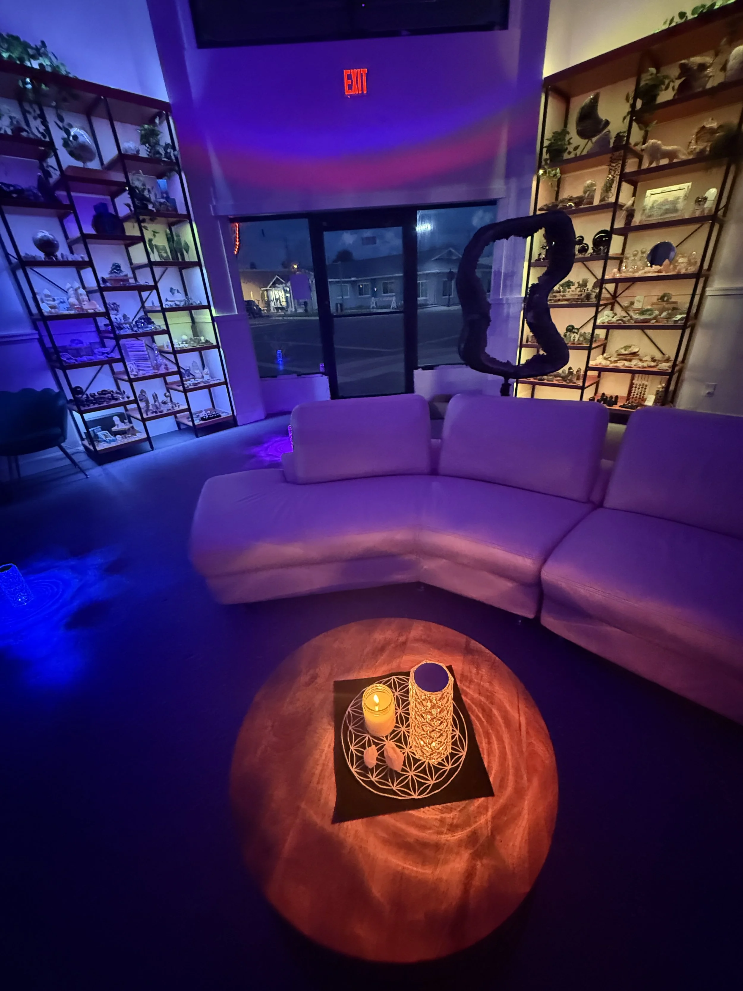 A cozy, dimly lit room with purple and blue lighting, featuring a white sectional sofa, a round wooden table with a candle and decorative holder, and two metal shelving units filled with ornaments and plants. A large window or glass door shows a street scene outside, with an illuminated exit sign overhead.