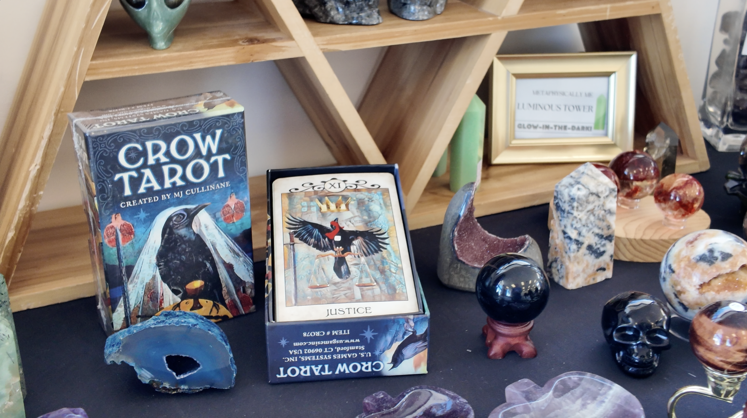 A display featuring tarot and or spiritual crystals, including a tarot deck titled 'Crow Tarot' by MJ Cullinan, and various polished stones, some carved into skulls and spheres, on a black surface with a wooden shelf background.