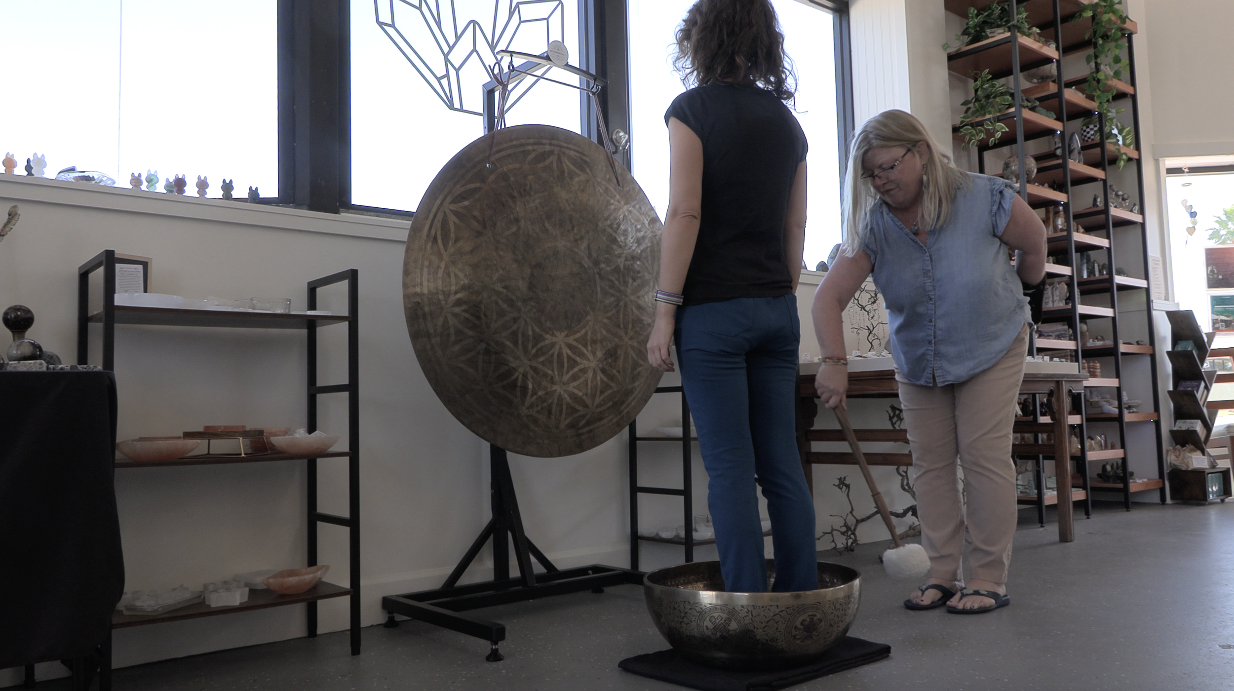 Two women in a store with shelves of pottery and decorations, one playing a large singing bowl with a mallet, near a large gong.
