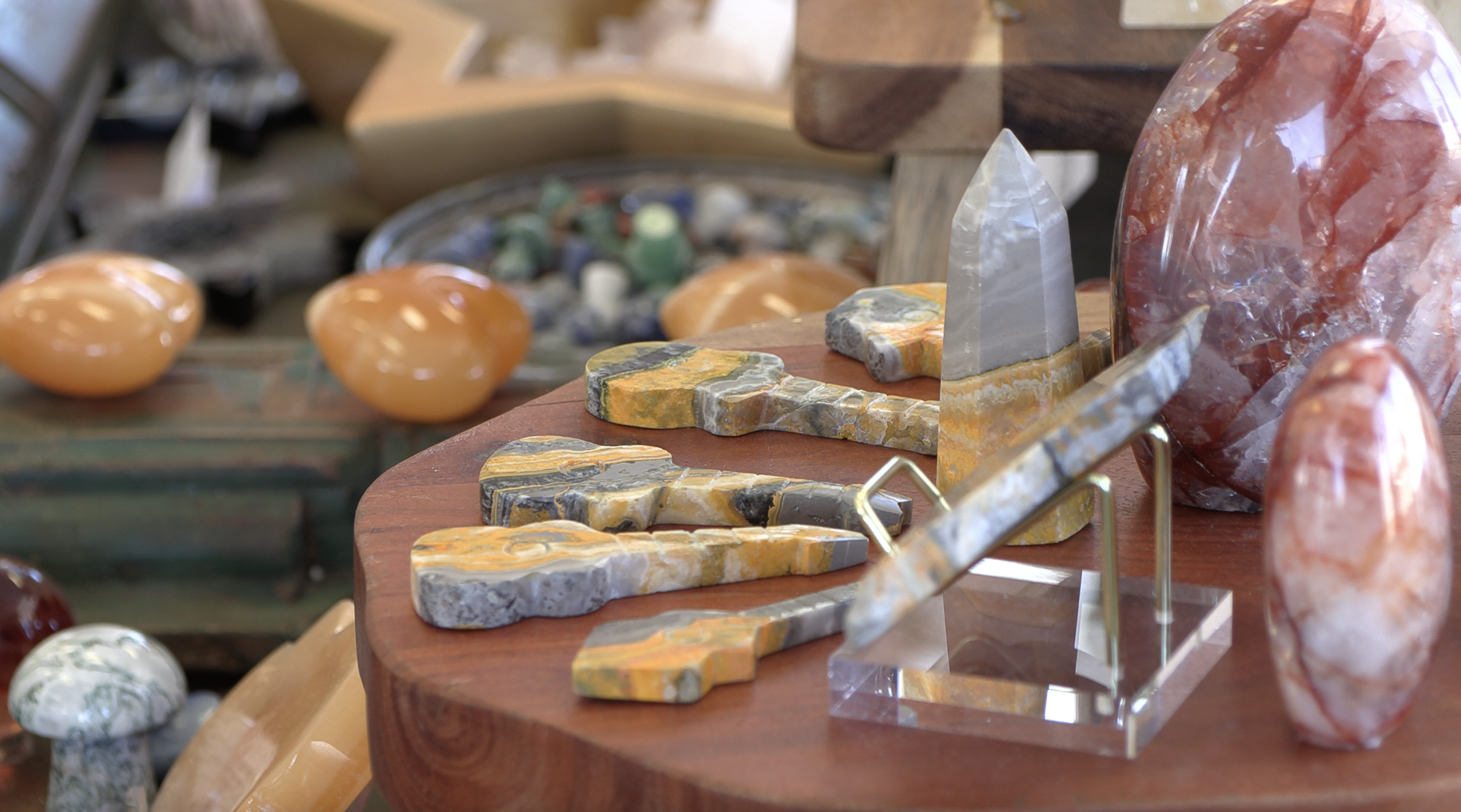 Collection of polished and raw gemstones and minerals, including a large red and orange stone and a gray crystal, displayed on a wooden table.