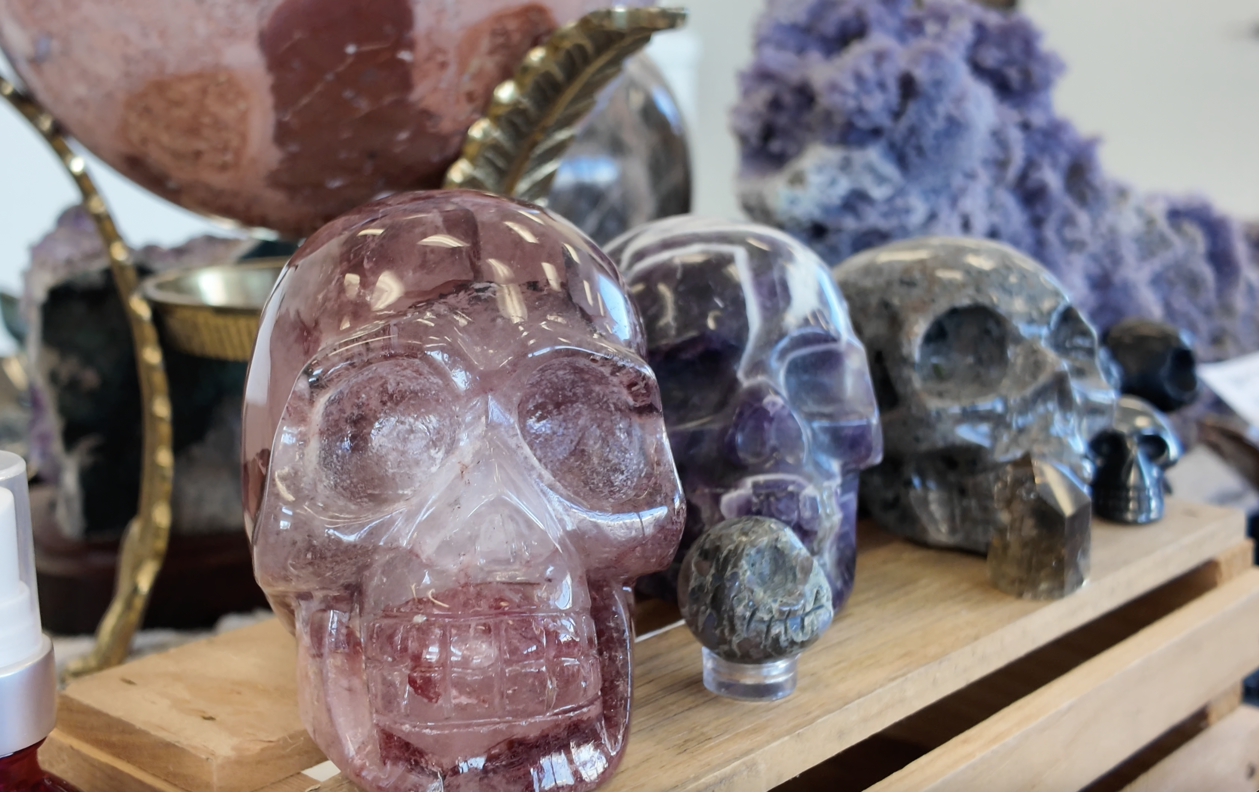 A collection of crystal skull sculptures, including a prominent pink one, displayed on a wooden shelf with purple mineral clusters in the background.