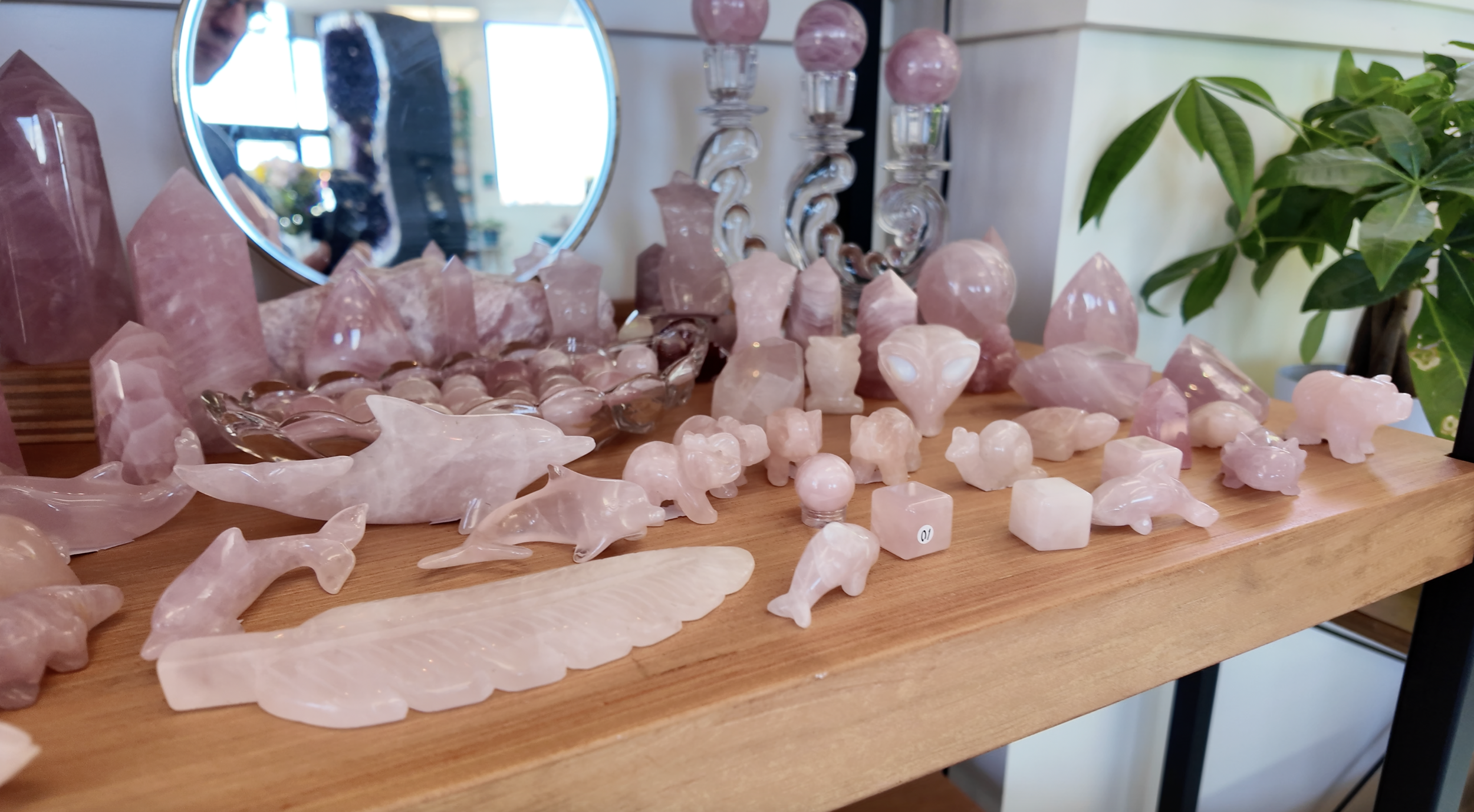 A wooden table displaying various pink and white rose quartz sculptures including animals, hearts, spheres, and geometric shapes, with a mirror, decorative glass candlesticks, and a plant in the background.