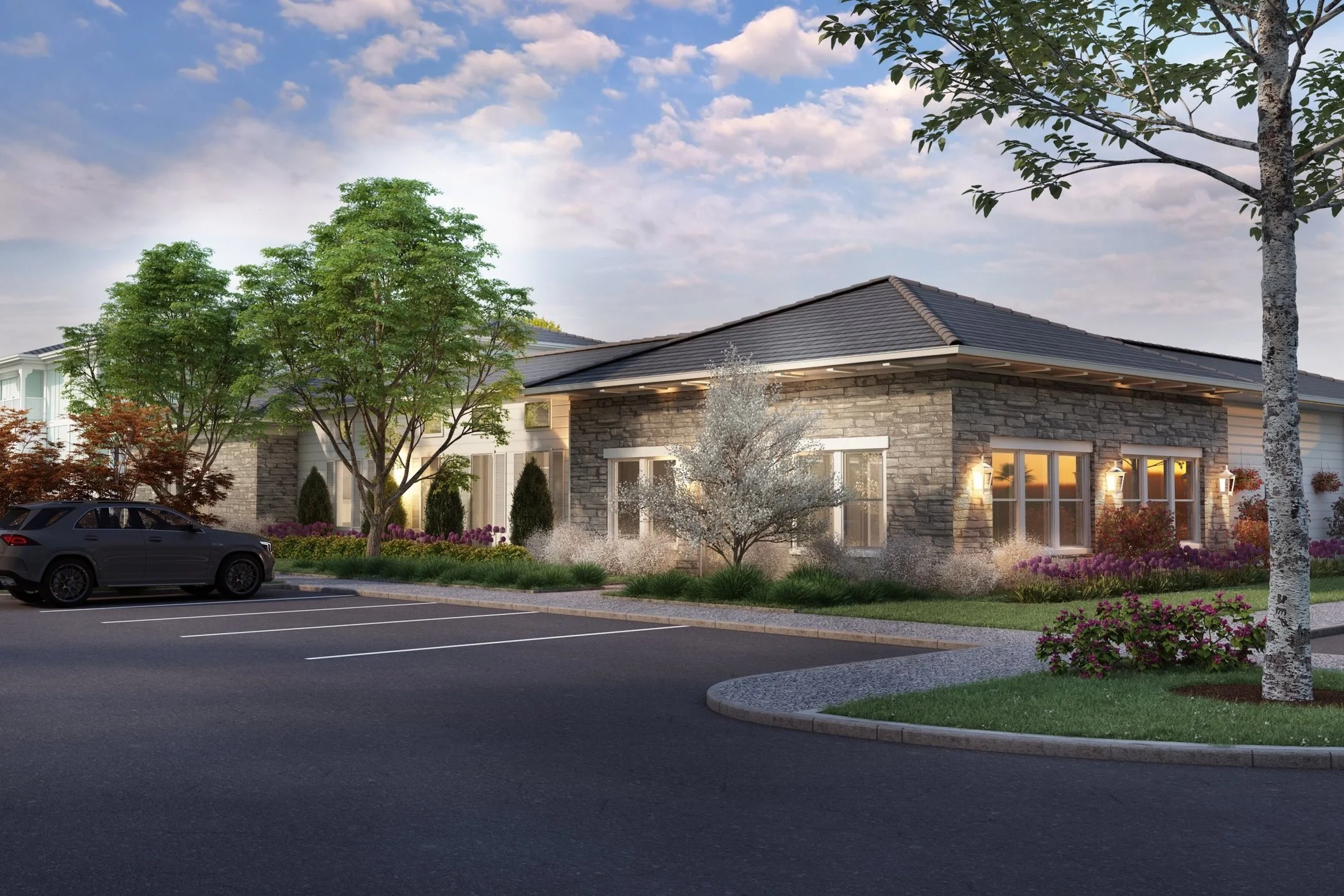 Residential building with stone exterior, landscaped garden with trees and flowers, parking lot with a black car, and a cloudy sky at dusk.