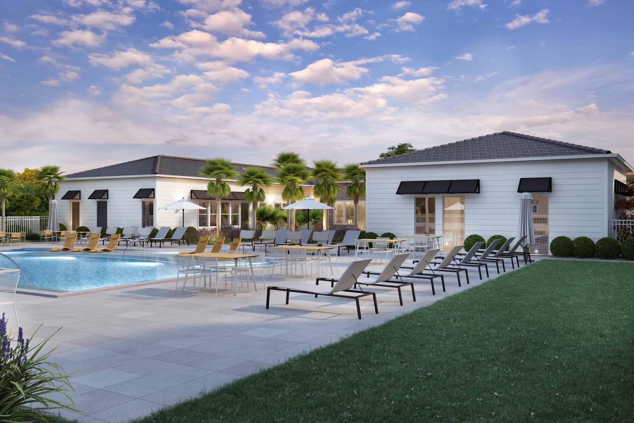 Residential swimming pool area with lounge chairs, tables with umbrellas, and white buildings with black awnings, surrounded by lush green grass and palm trees under a partly cloudy sky.