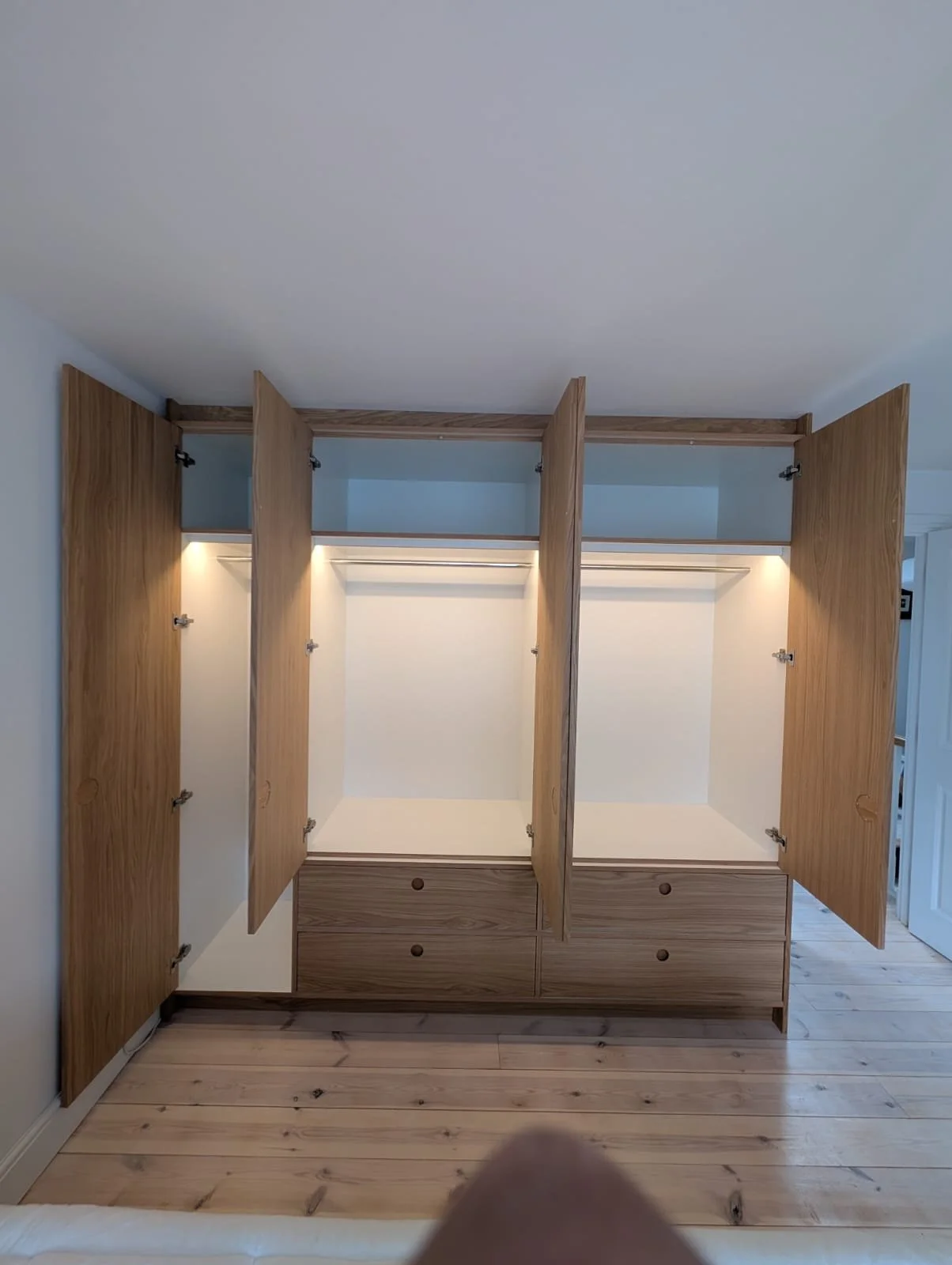 Open empty wooden wardrobe with multiple compartments and drawers, built-in lighting, in a room with light-colored wooden floor.