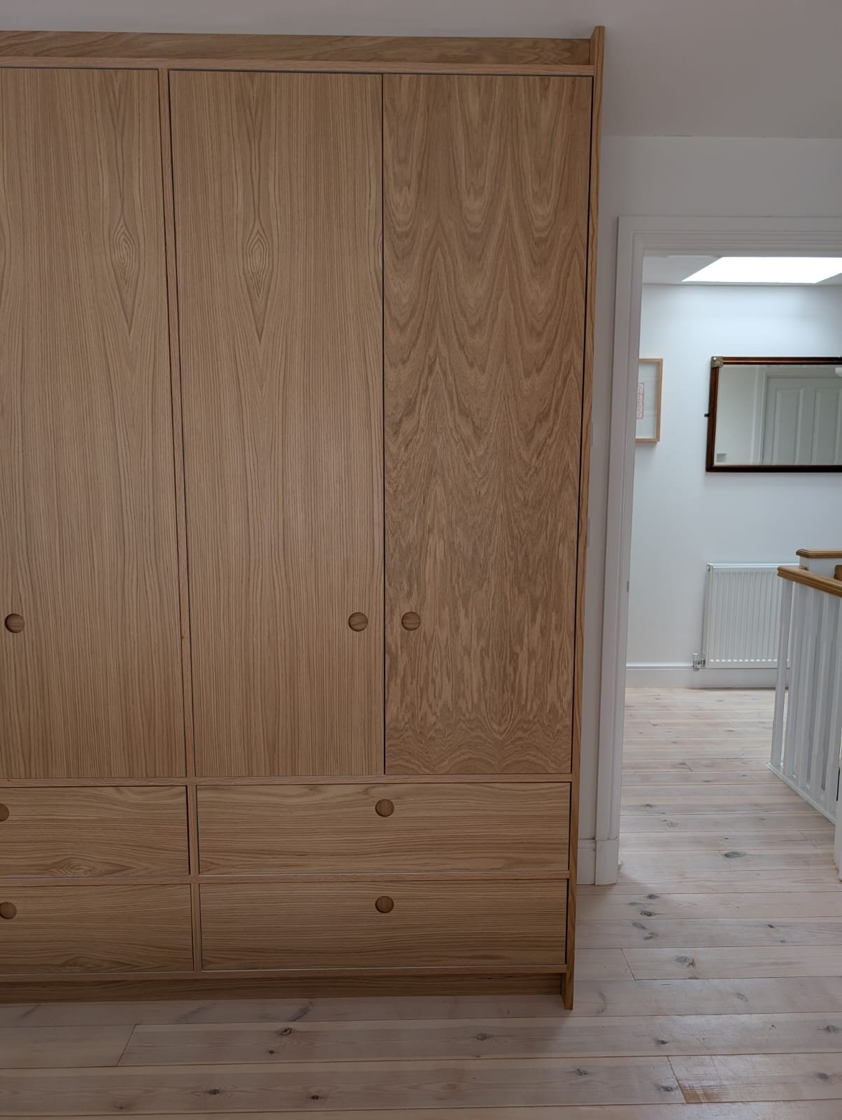 Wooden wardrobe with doors and drawers, located next to a door opening into a brightly lit room with white walls and hardwood floors.