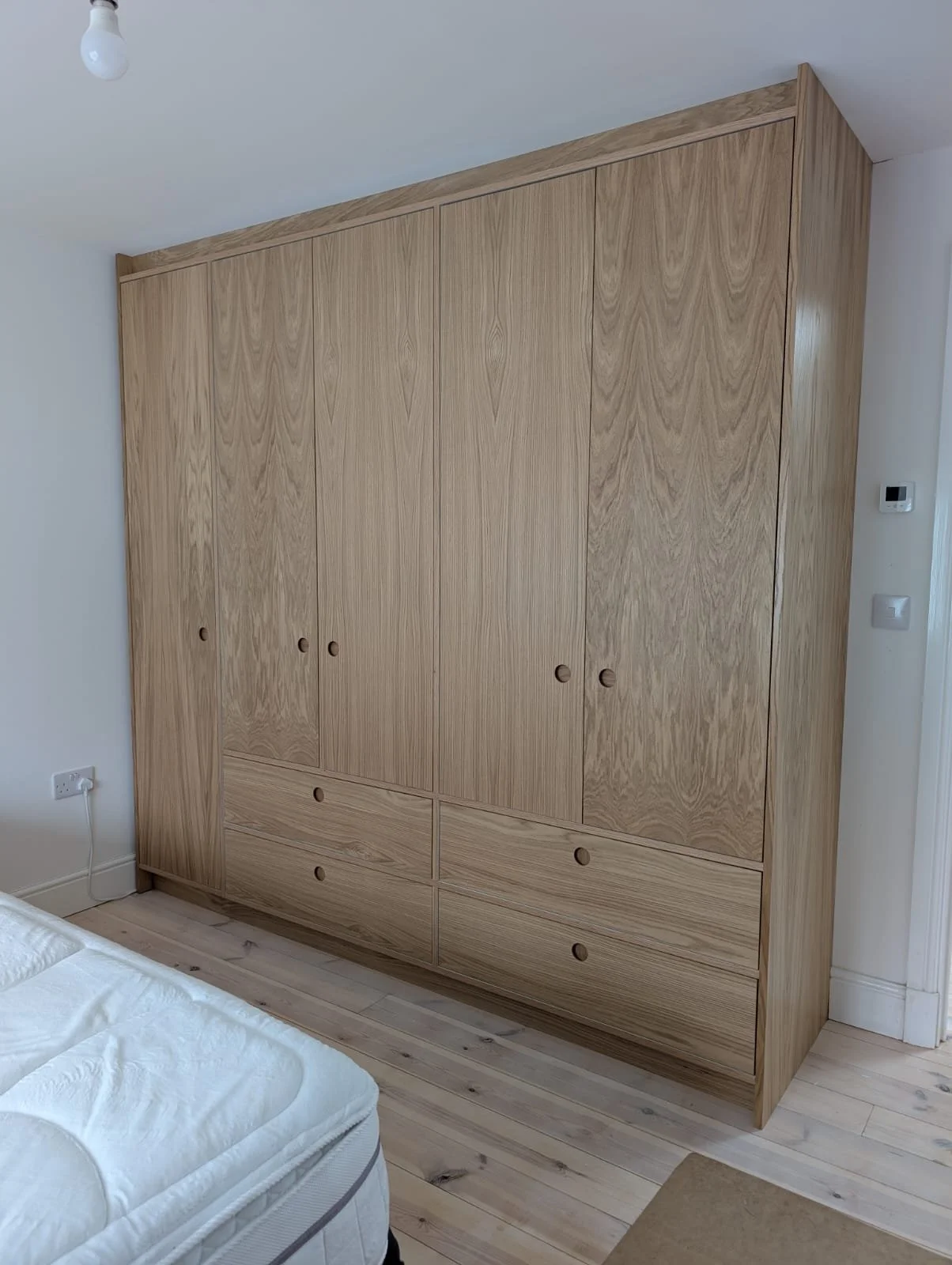 A large wooden wardrobe with four doors and four drawers, located in a room with light-colored wooden flooring and white walls.