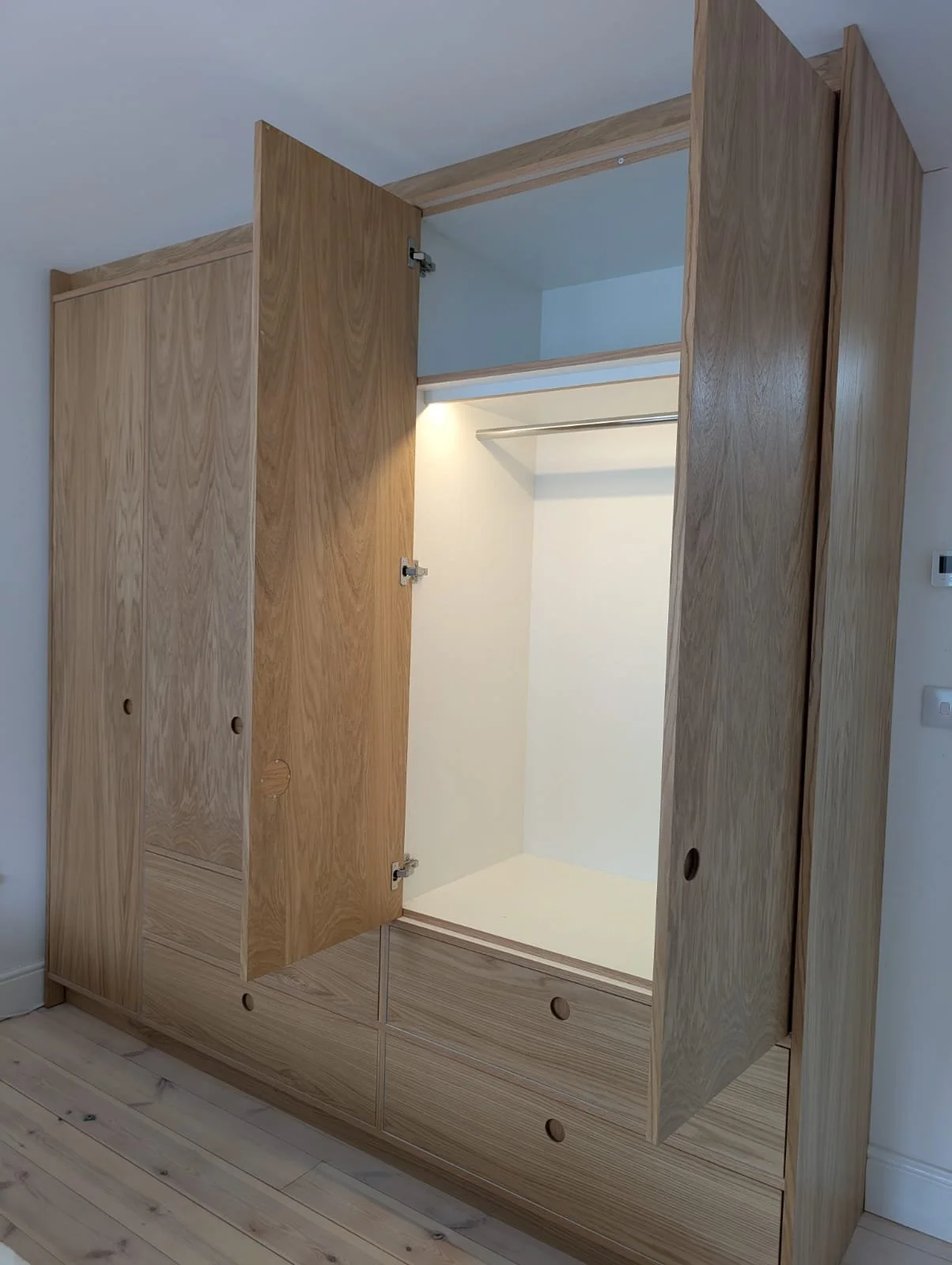 An empty wooden wardrobe with open doors, showing the interior with a hanging rod and white interior walls, placed against a white wall with a light wooden floor.