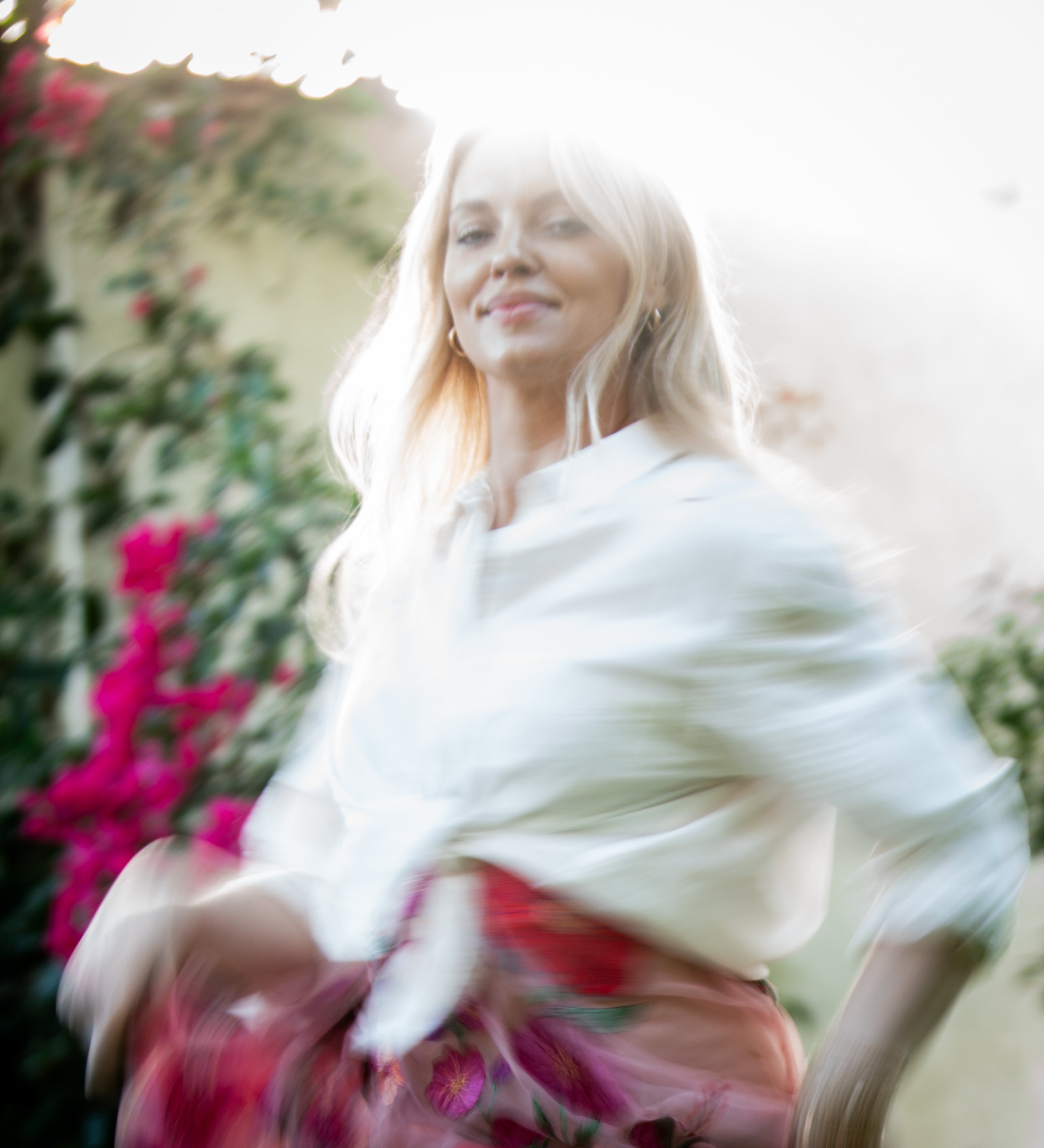 Close-up of a woman with blonde hair and earrings, smiling, with a blurred background and a large leafy plant on the side. The image has a bright, overexposed lighting.
