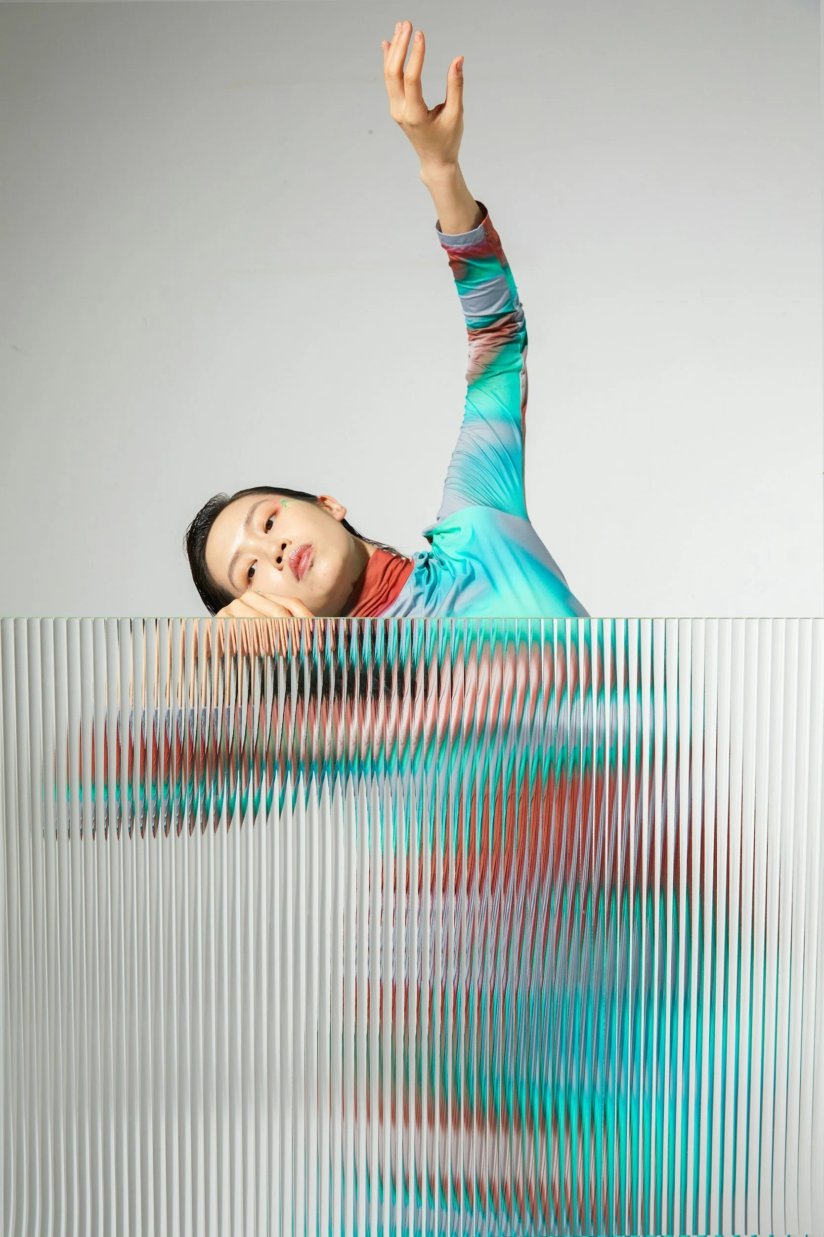 A woman with long dark hair lying on her side with her head resting on her arm, wearing a colorful long-sleeved shirt, with her eyes looking at the camera and her other arm raised, reaching upward. The background is plain and gray, and there is a decorative element at the bottom of the image.