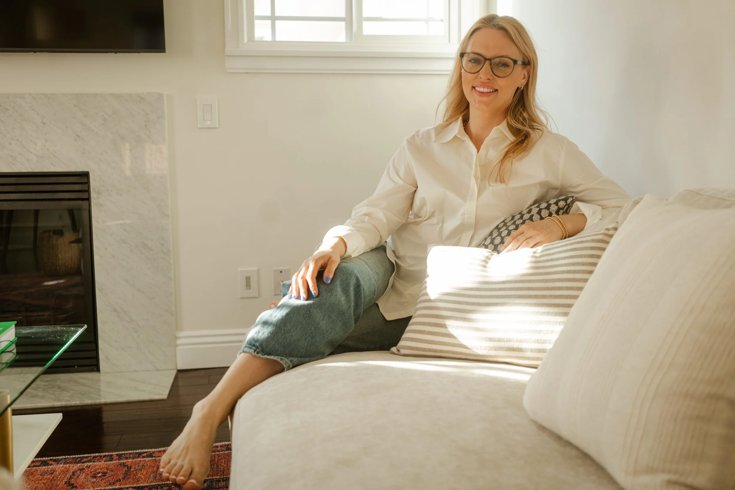 A woman with blonde hair, glasses, and a bright smile, sitting on a beige sofa in a sunlit living room, wearing a white blouse and gray pants, with a decorative pillow on the sofa.