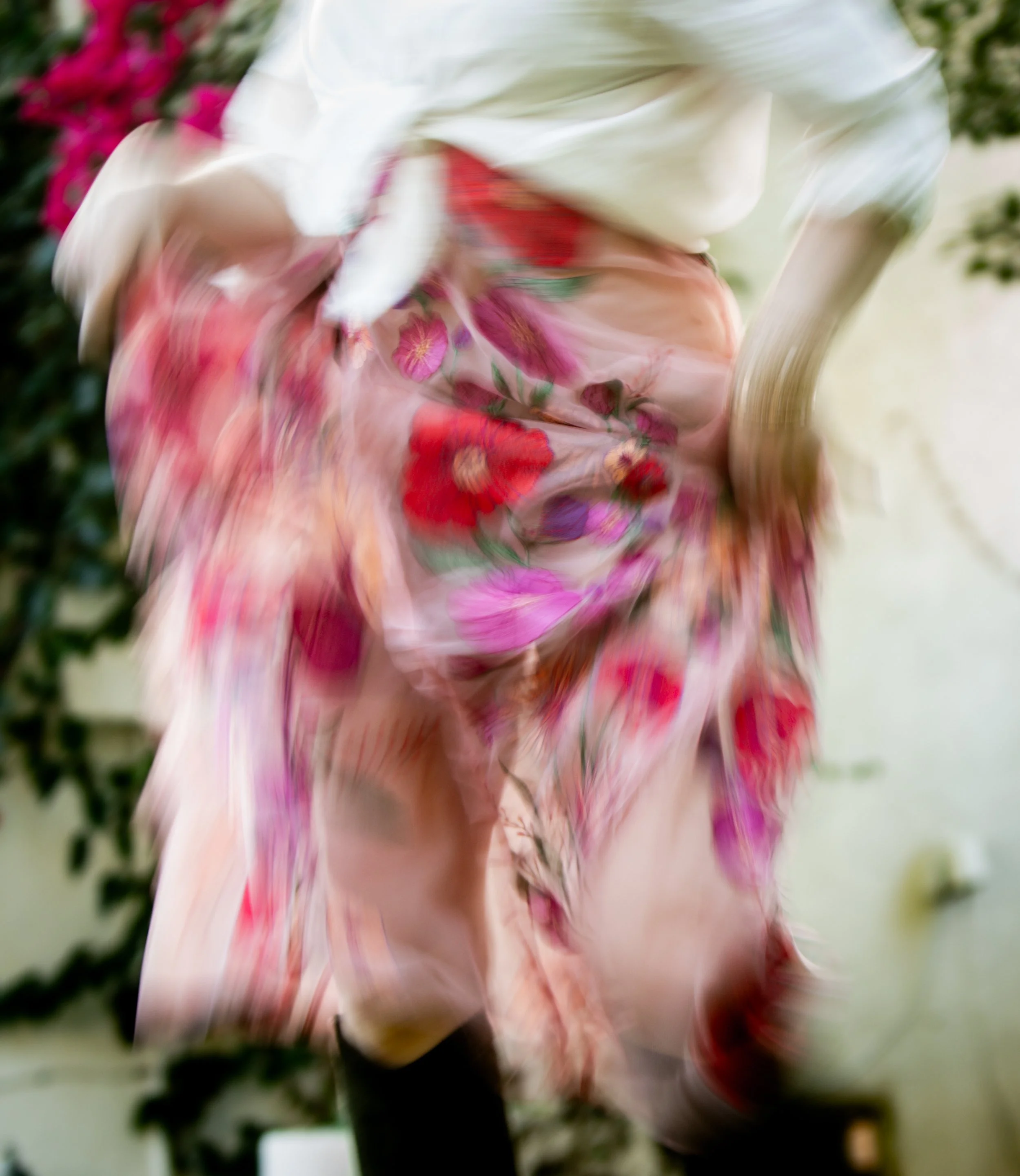 Blurry photograph of a person wearing a floral dress, with pink and red flowers, and a white shirt, standing in front of green foliage.