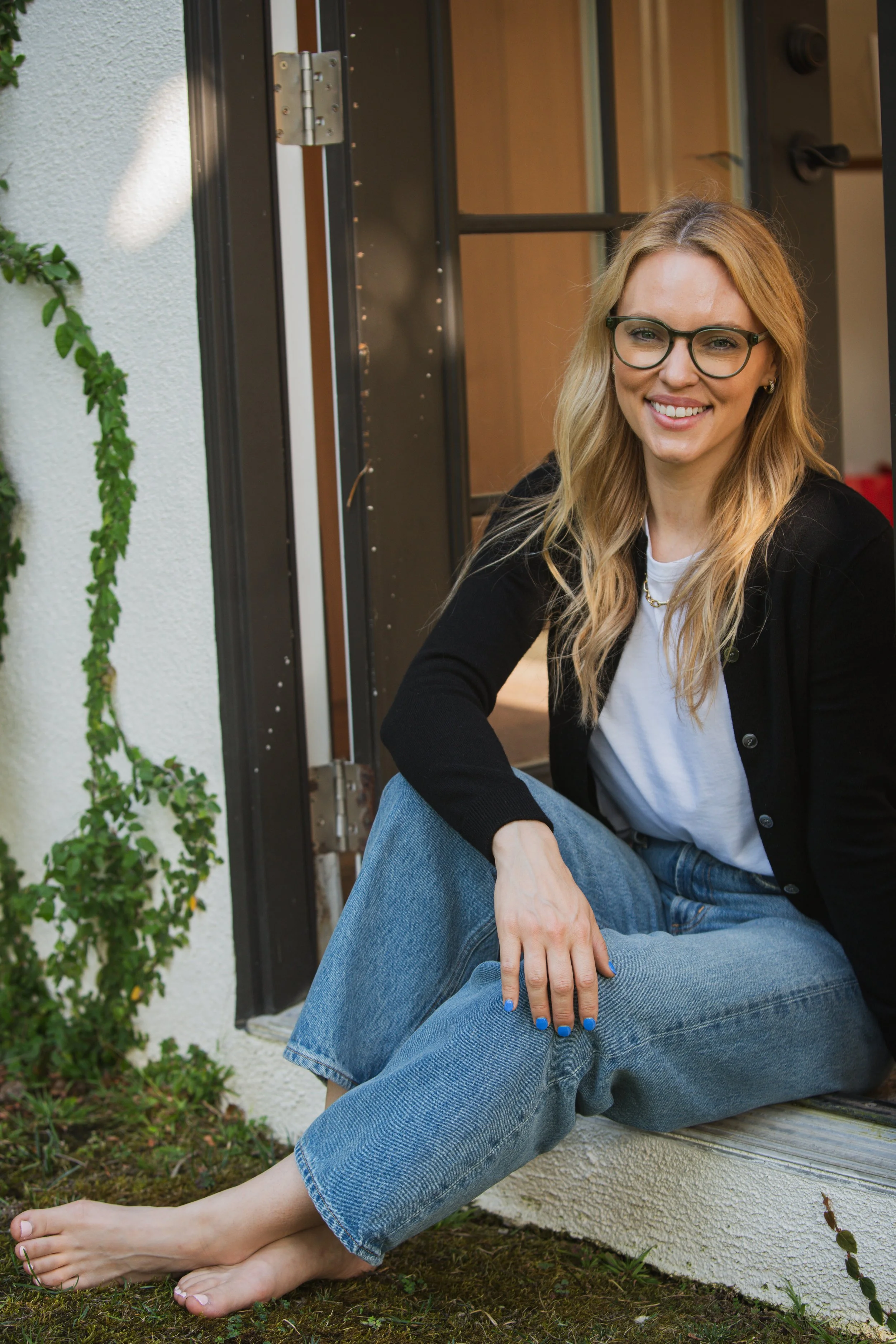 A young woman with blonde hair, glasses, and a black jacket sitting on a porch step, smiling, with her legs crossed at the ankle, barefoot, next to a white wall with greenery.