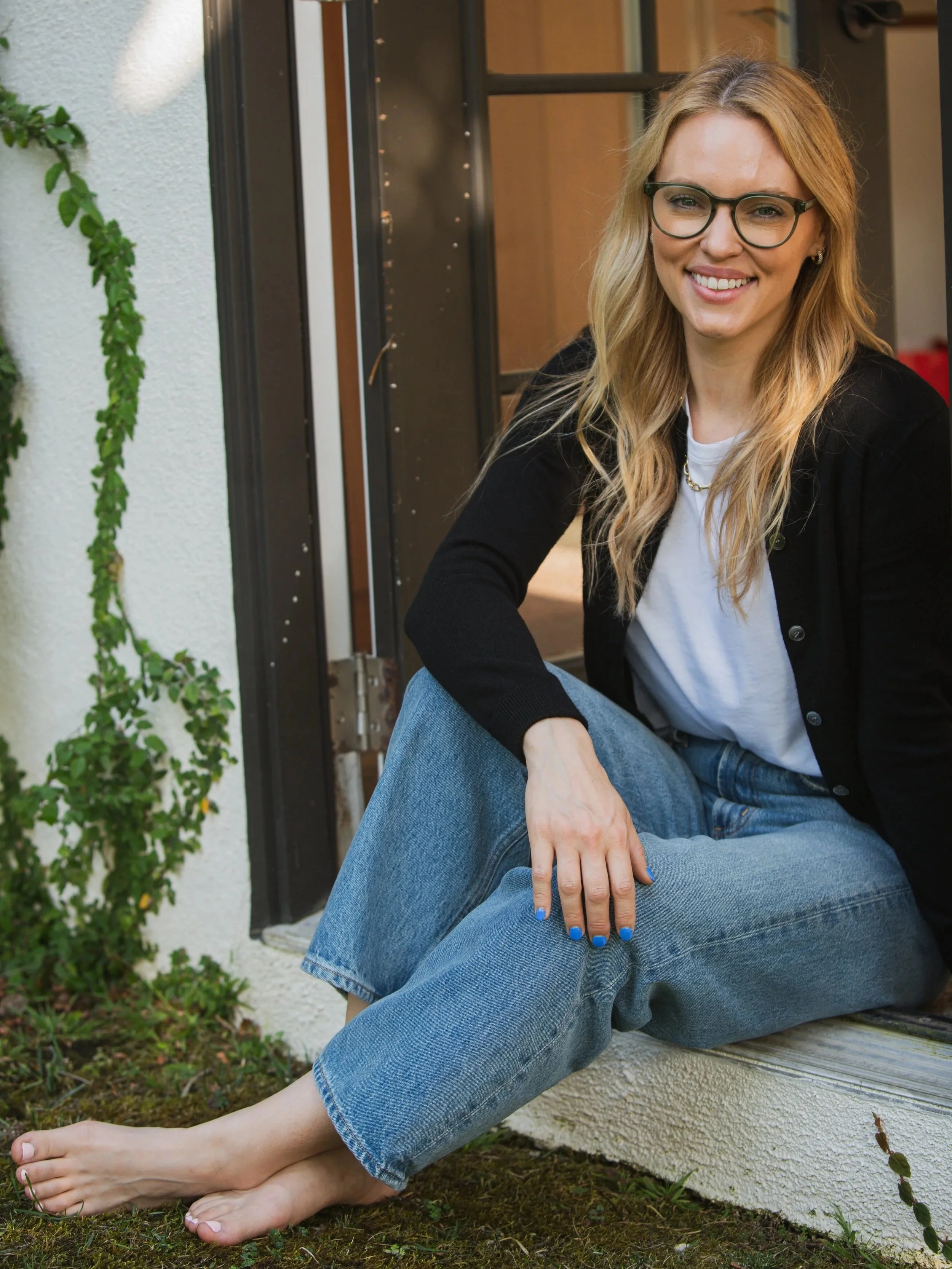 A woman wearing glasses, a black cardigan, and blue jeans sitting outside next to a wall with a plant growing nearby. She is smiling and has her legs crossed, with her feet bare on the ground.