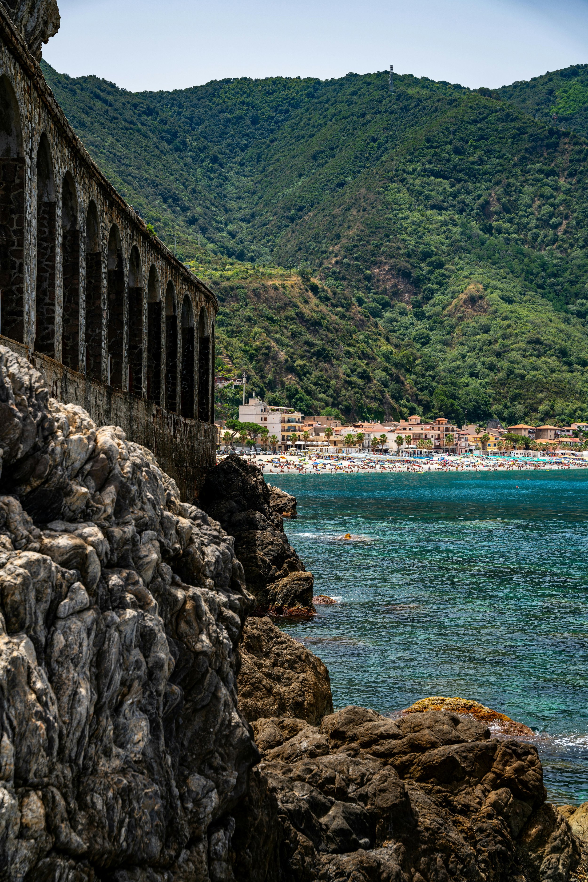 A rocky shoreline with a historic stone and arch aqueduct along the coast, overlooking a beach with umbrellas and a town nestled among lush green mountains under a blue sky.
