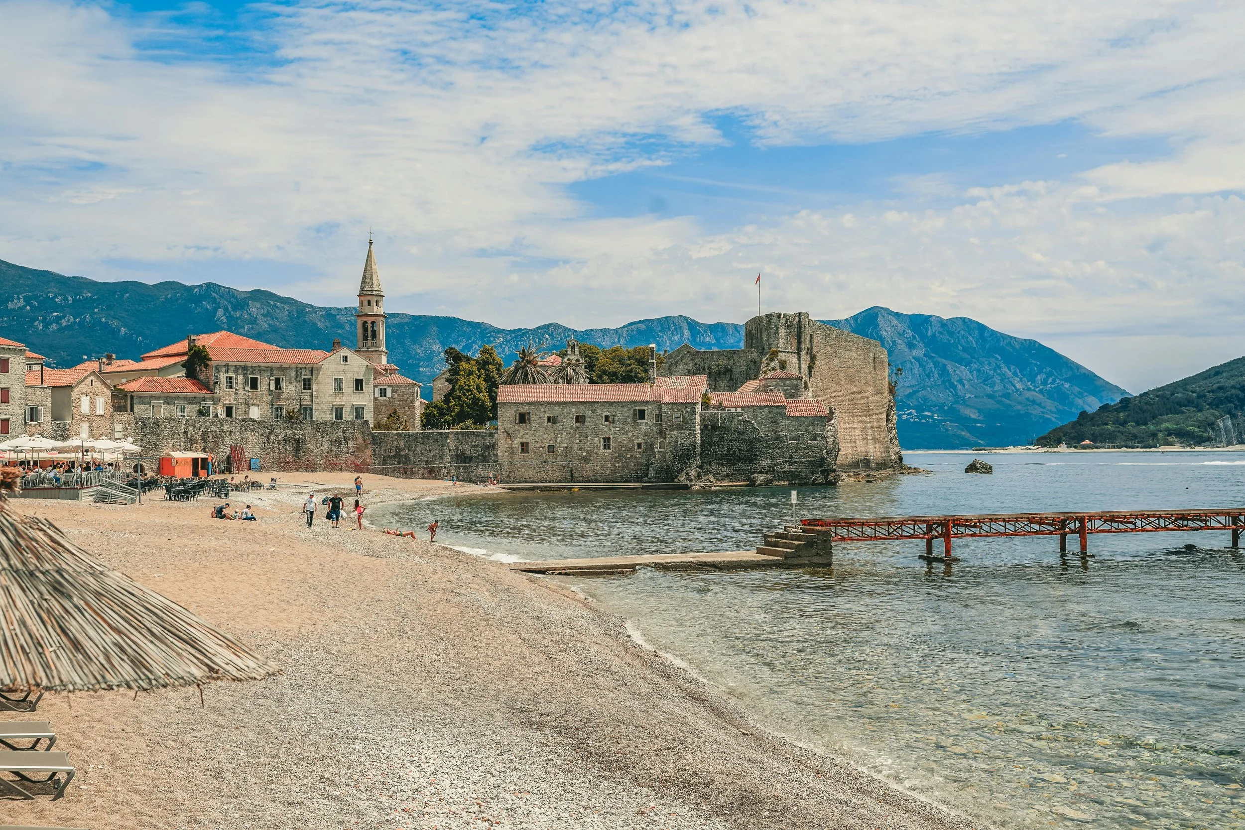 A beach with sand and a thatched umbrella in the foreground, people walking along the shore, historic stone buildings and a castle on a peninsula, with mountains in the background under a partly cloudy sky.
