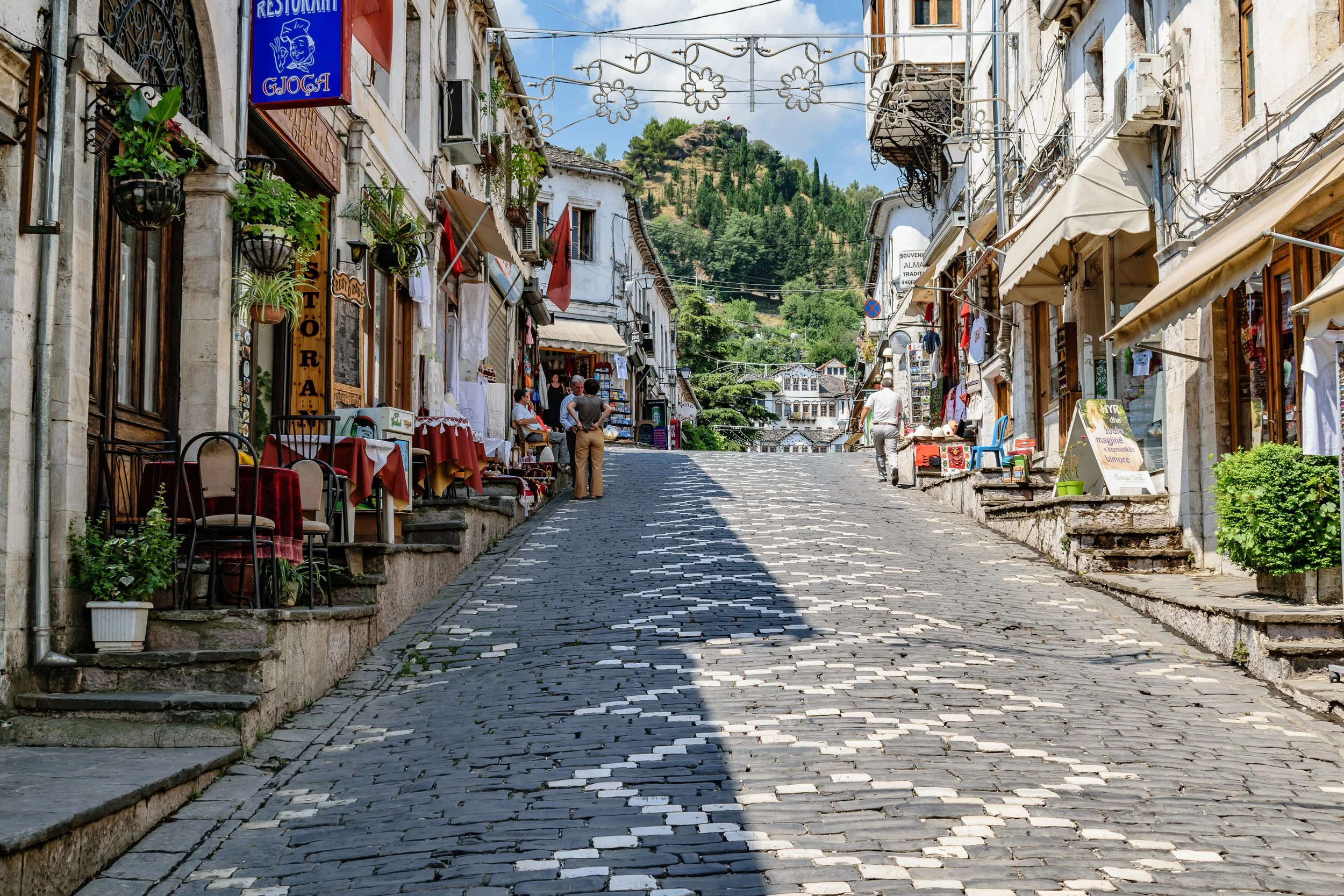 A cobblestone street in a European village with small shops and cafes on both sides. Some people are walking and browsing. Buildings have awnings, balconies with plants, and signs. A hill with trees is visible in the background.