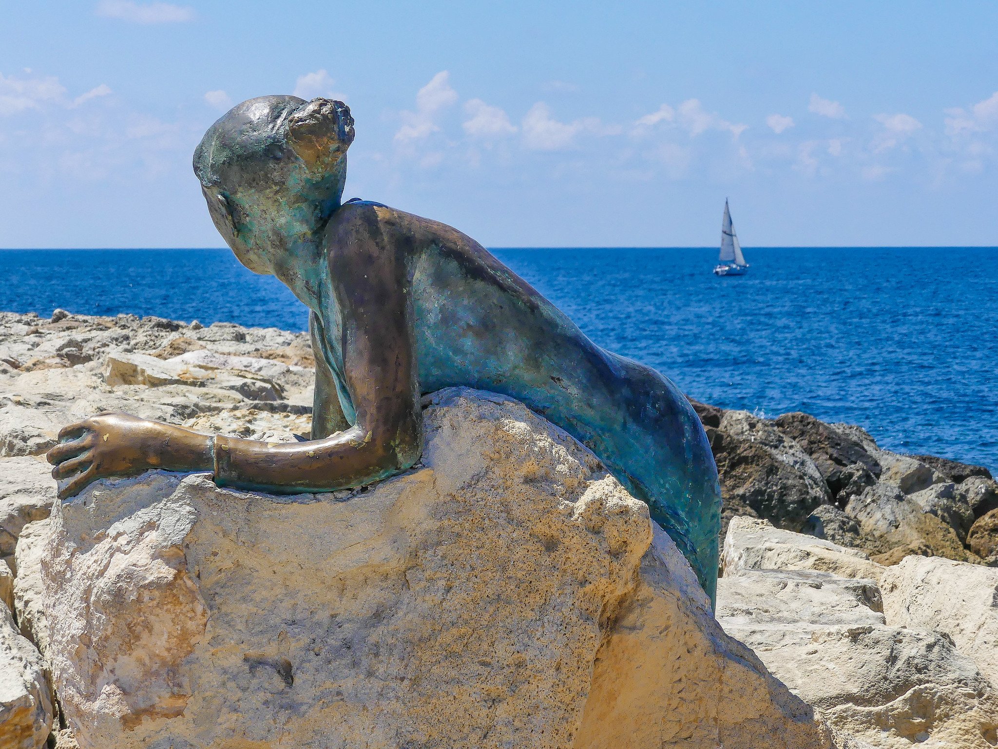 Bronze sculpture of a woman lying on rocks by the sea, with a sailboat in the background