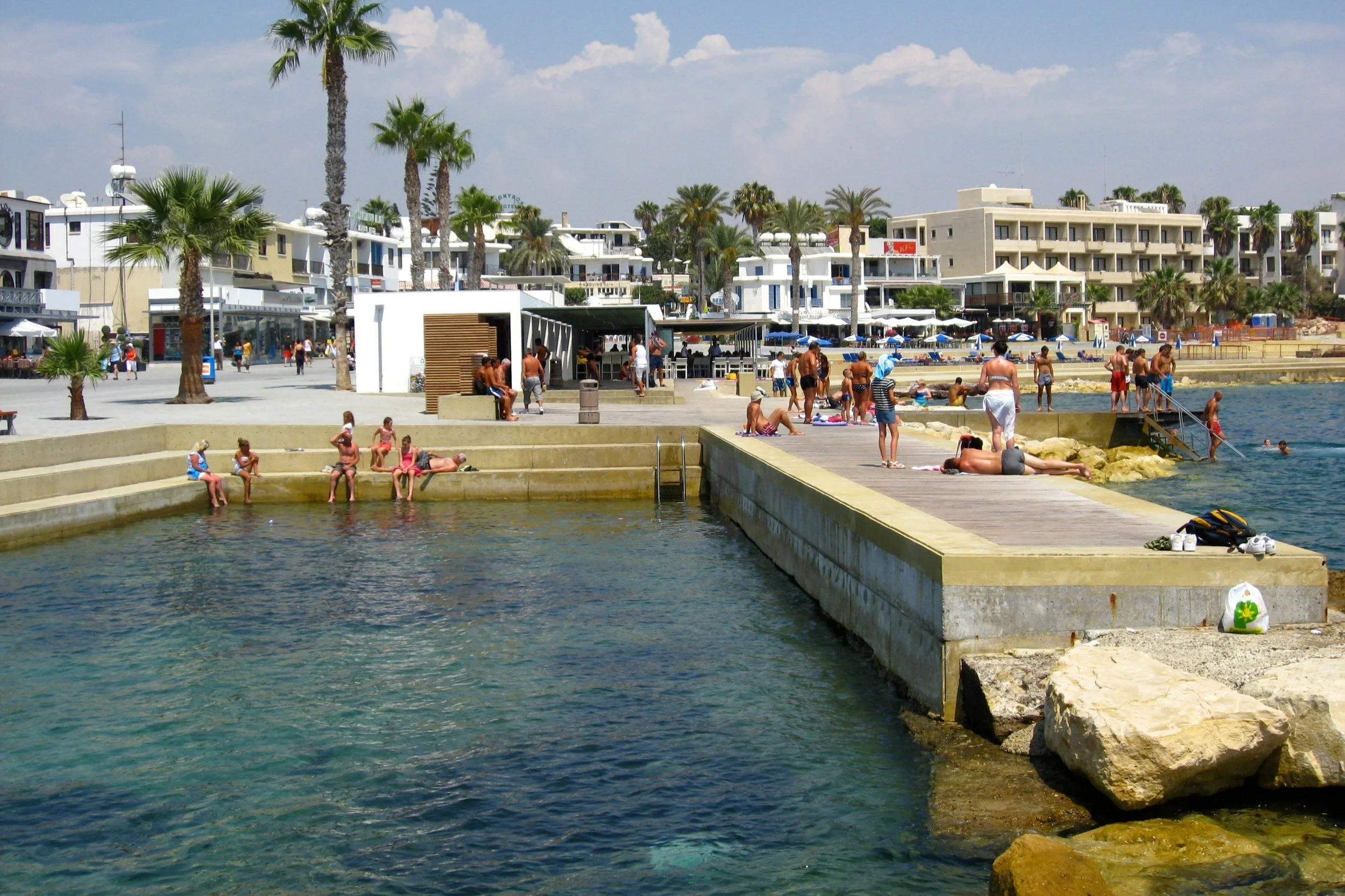 Beachside scene with people relaxing on a concrete pier, swimming, and sunbathing near palm trees and modern white buildings under a partly cloudy sky.