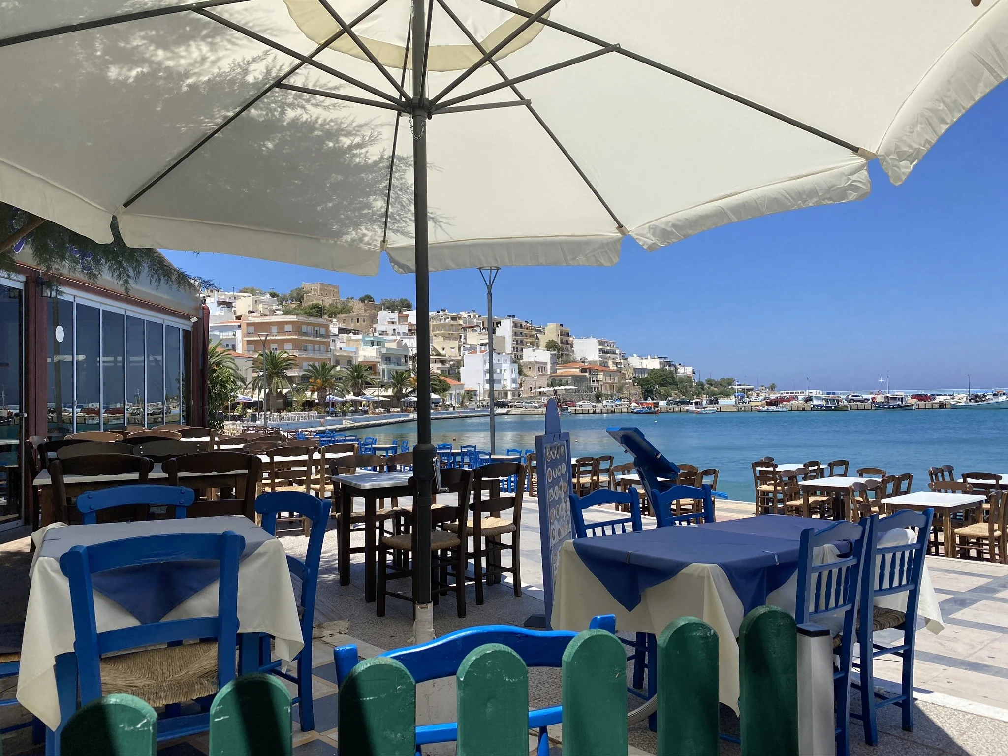 Outdoor seaside restaurant with blue chairs and tables covered with white and blue tablecloths, large white umbrella, view of boats and hillside buildings across the water.
