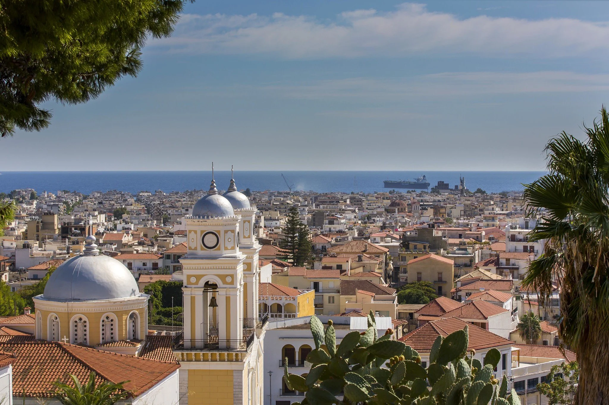 Cityscape of a coastal town with churches, trees, and a view of the ocean with ships in the distance.