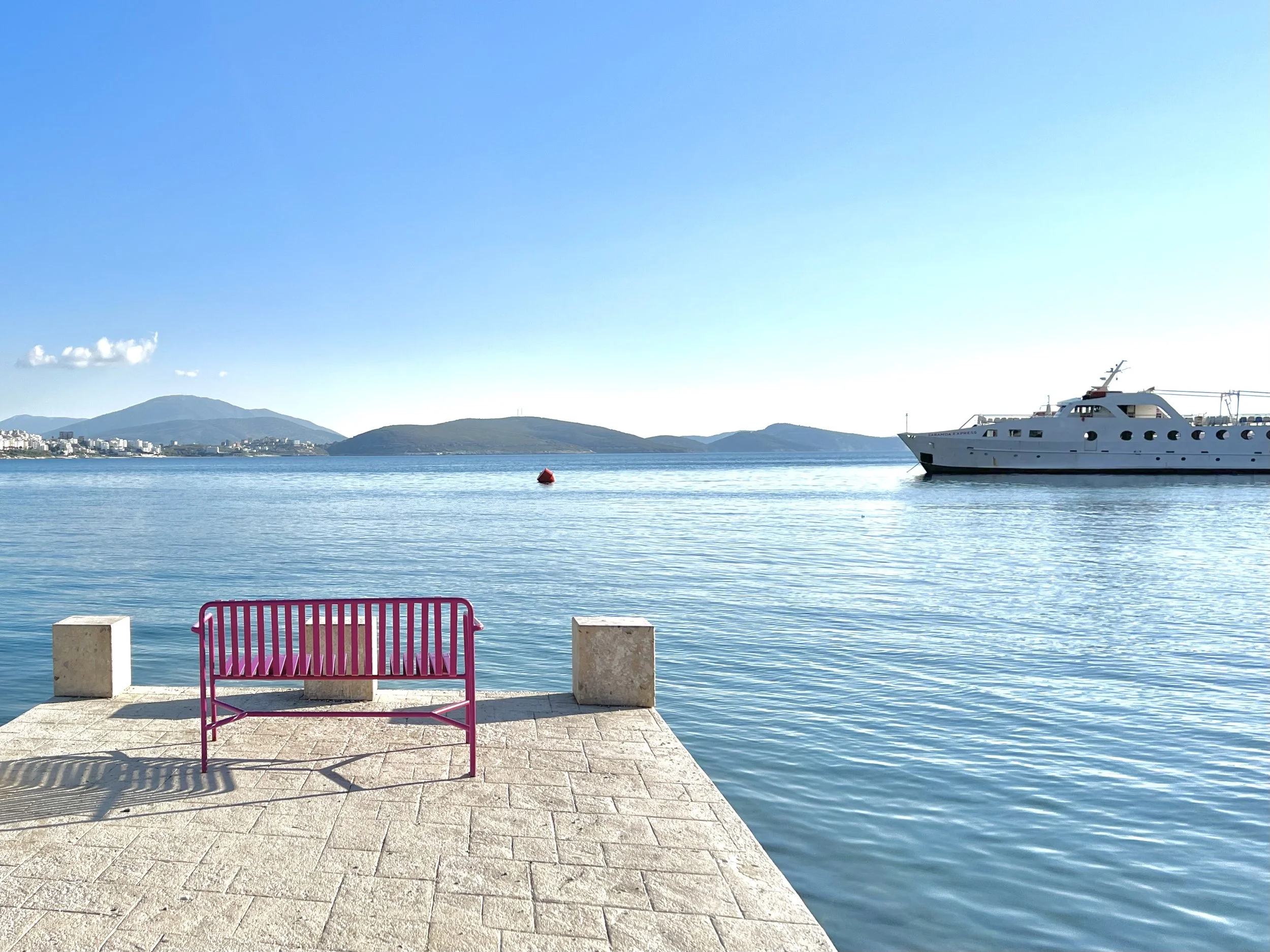 A pink bench on a stone pier overlooking a calm body of water with a large white yacht to the right and an island or peninsula in the distance under a clear blue sky.
