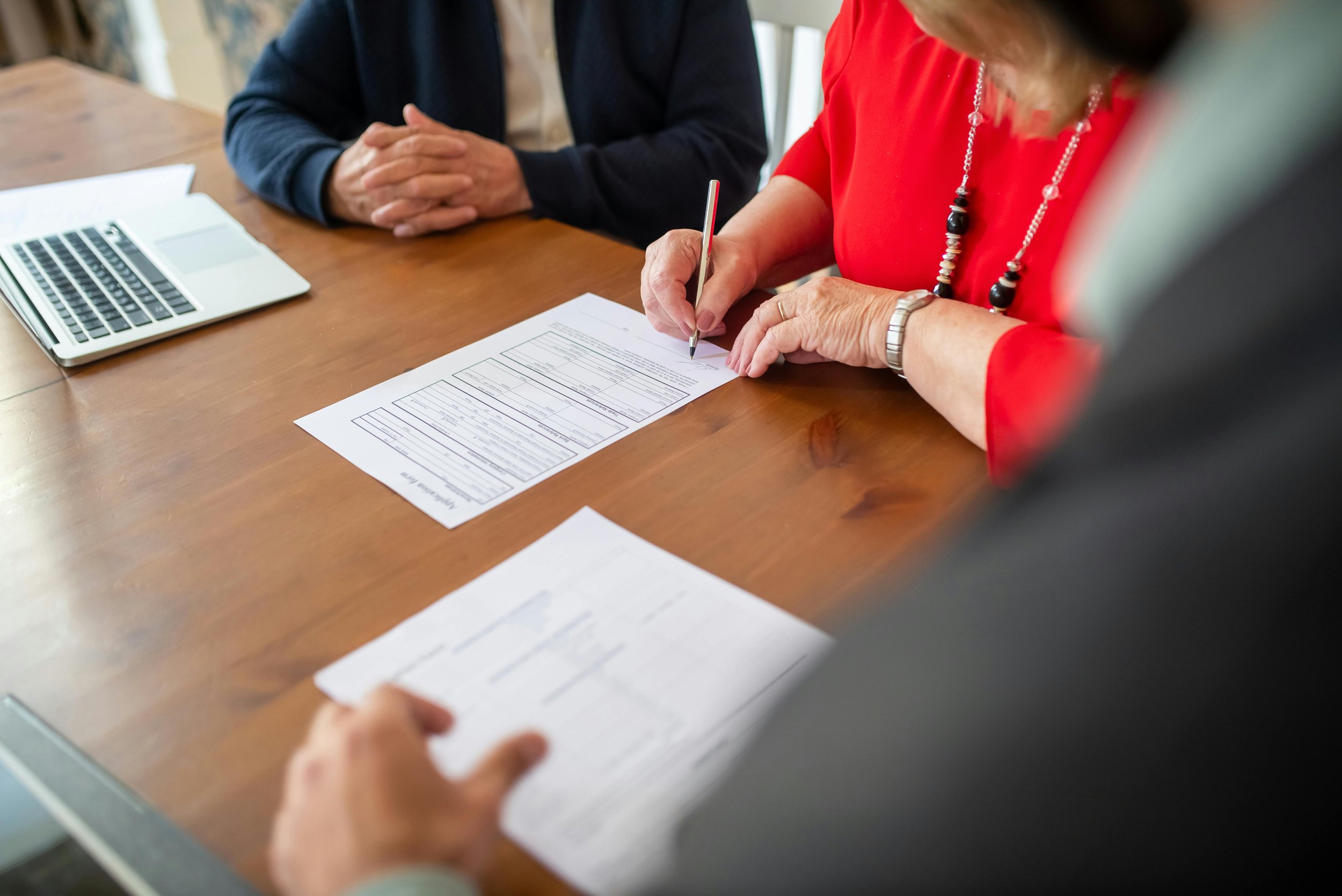 Person reviewing and signing documents at a wooden table with a laptop nearby.