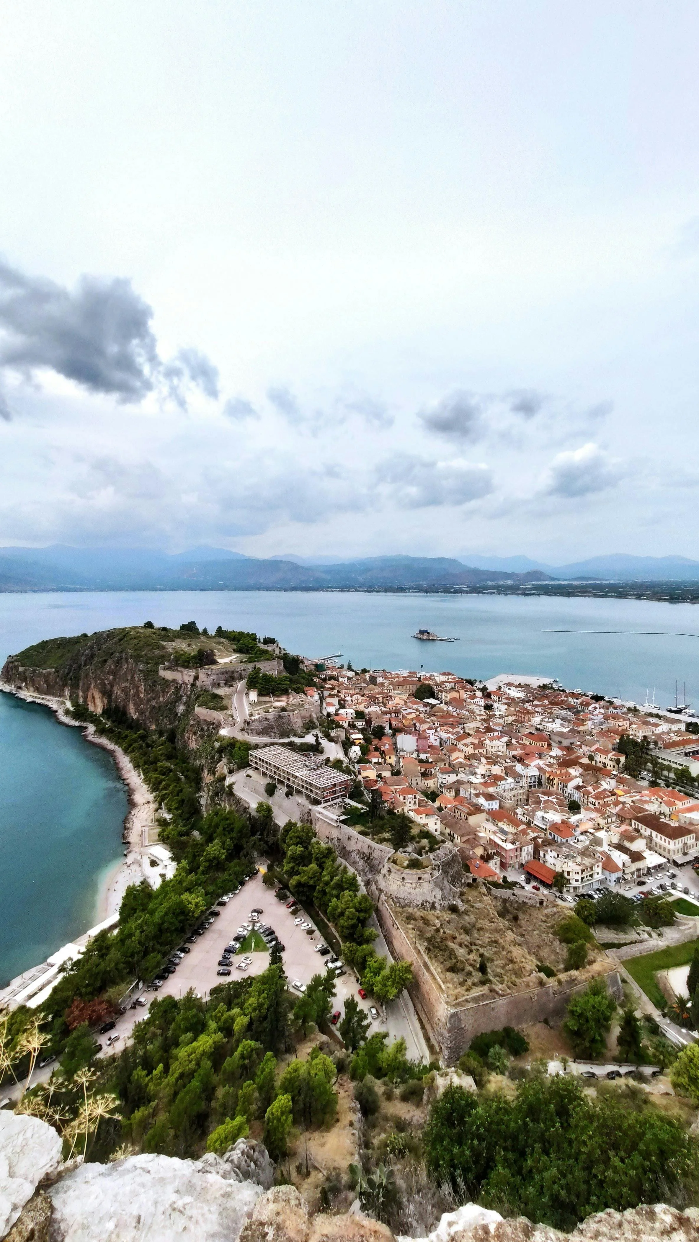 Aerial view of a coastal town with a castle wall, parking lot, and a beach, overlooking the sea and mountains in the distance.
