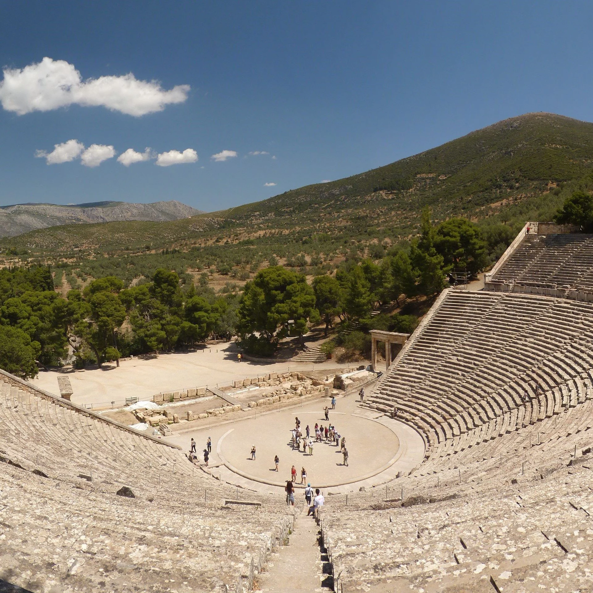 Ancient Greek open-air theater with stone seating, a stage area, and a group of tourists exploring, set in a mountainous landscape under a clear blue sky.