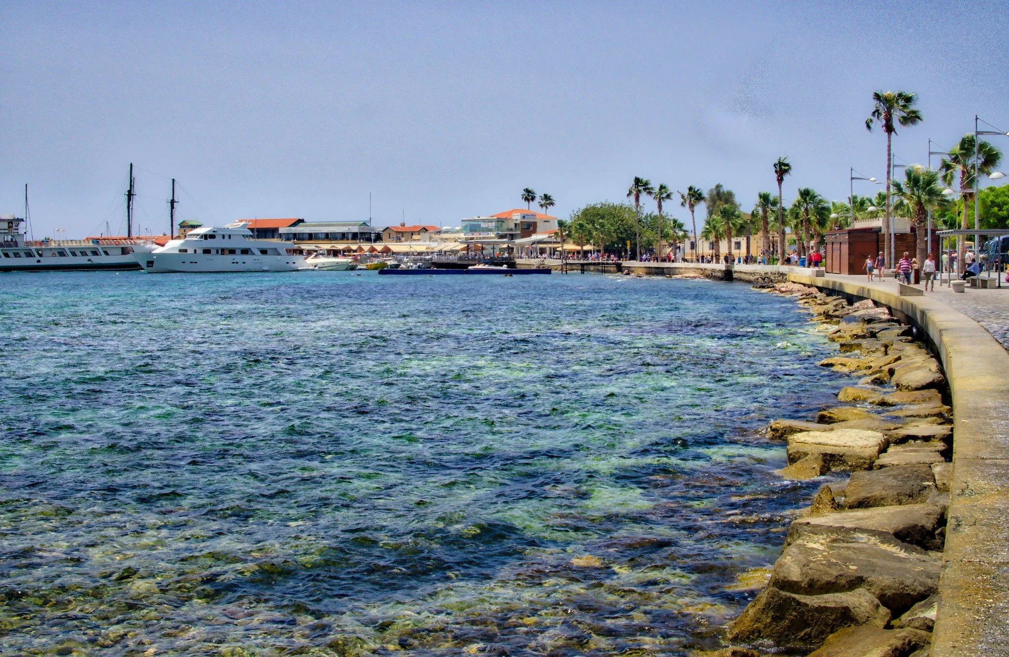 A coastal scene with a clear blue sky, calm water, boats docked at a marina, a walkway lined with palm trees, and people walking along the shore.