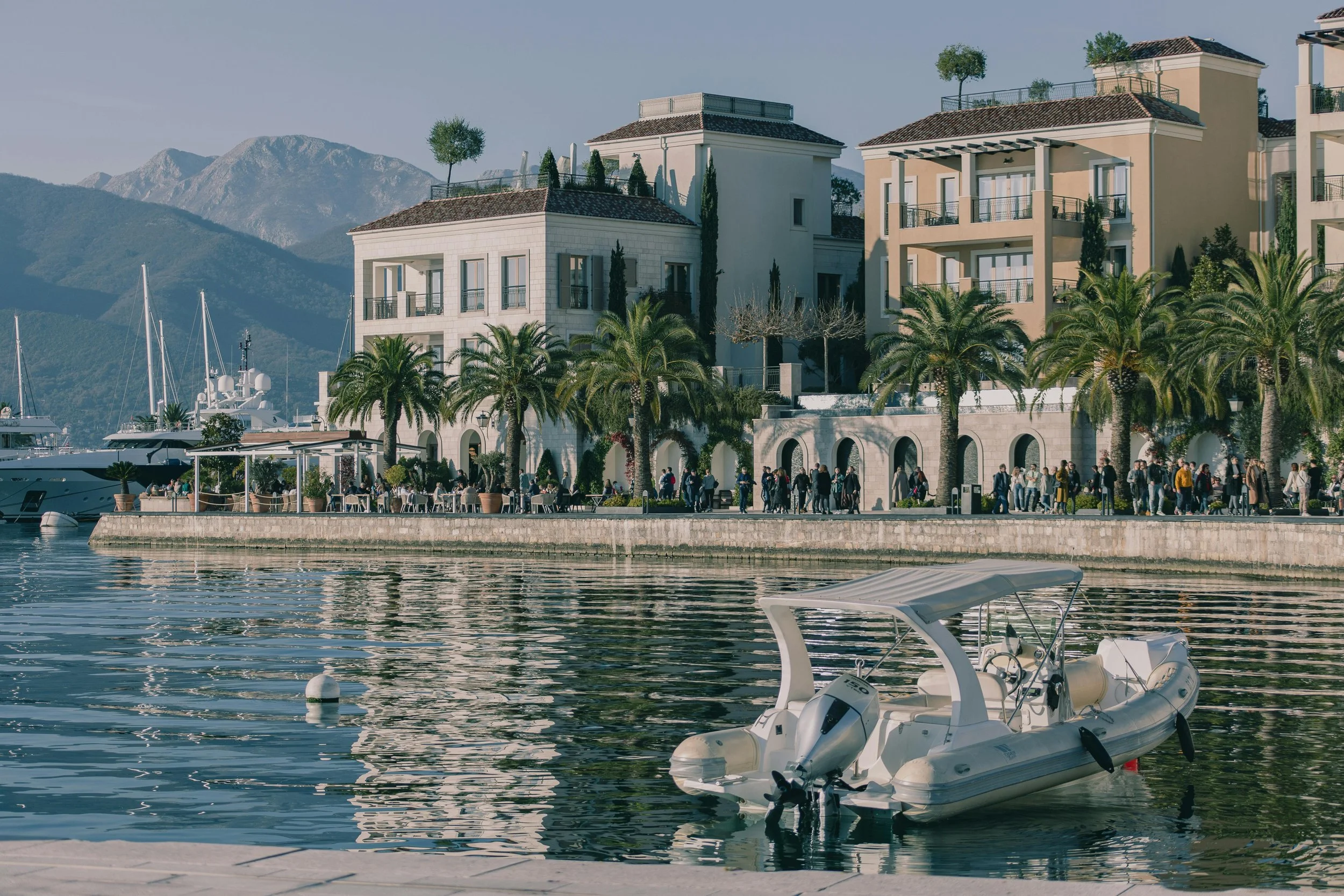 A waterfront scene with a small motorboat floating on calm water, lined with palm trees and a promenade. In the background, there are multi-story residential buildings and mountains under a clear sky.
