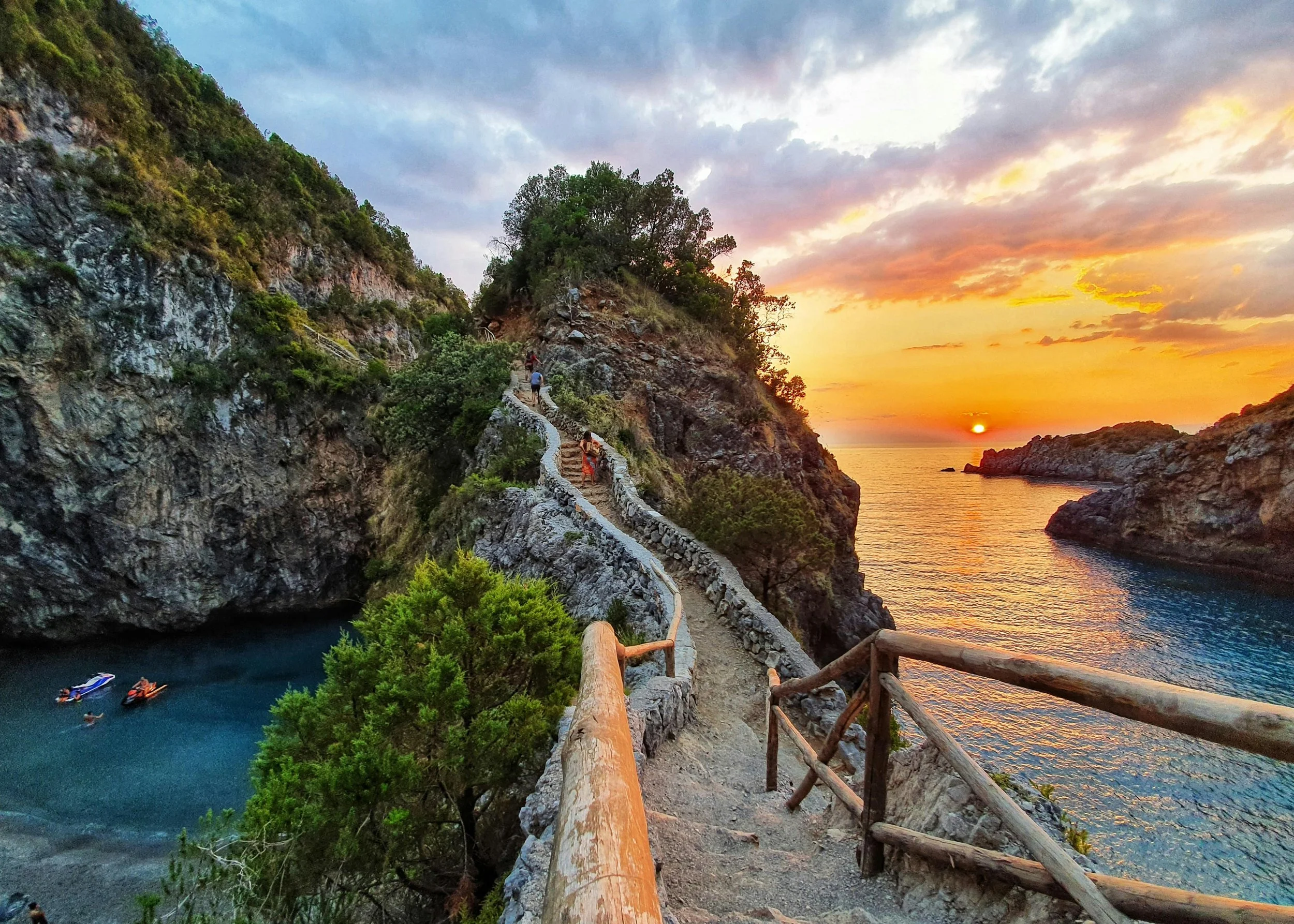 A narrow coastal trail with wooden railings winding along a rocky hillside overlooking the ocean during sunset. The sky is filled with colorful clouds, and a few people walk or sit on the trail. The ocean has a small cove with boats, and the sun is setting over the horizon.