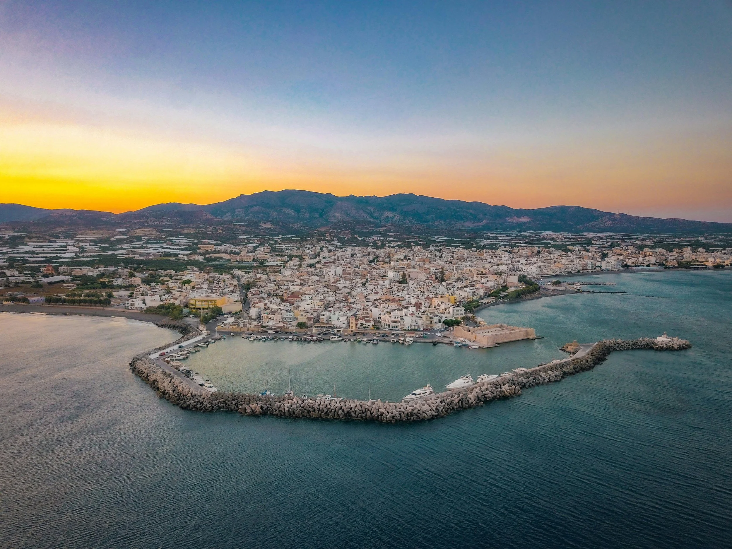 Aerial view of a coastal city at sunset with a harbor enclosed by a stone breakwater, a coastline, and a mountain range in the background.